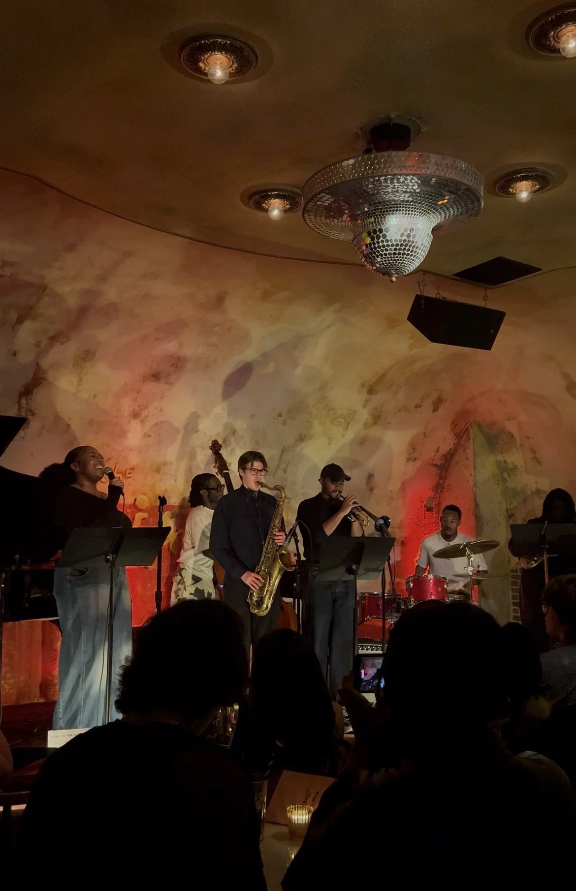 Jazz band performing on stage with musicians playing instruments, and a singer with a microphone, in a cozy venue with warm lighting and artistic wall mural.