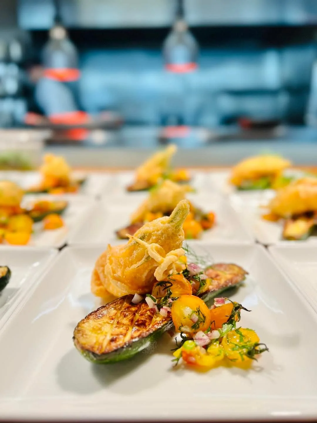 Gourmet dish with roasted zucchini, fried artichoke, and a colorful tomato salad on a white plate in a restaurant setting.