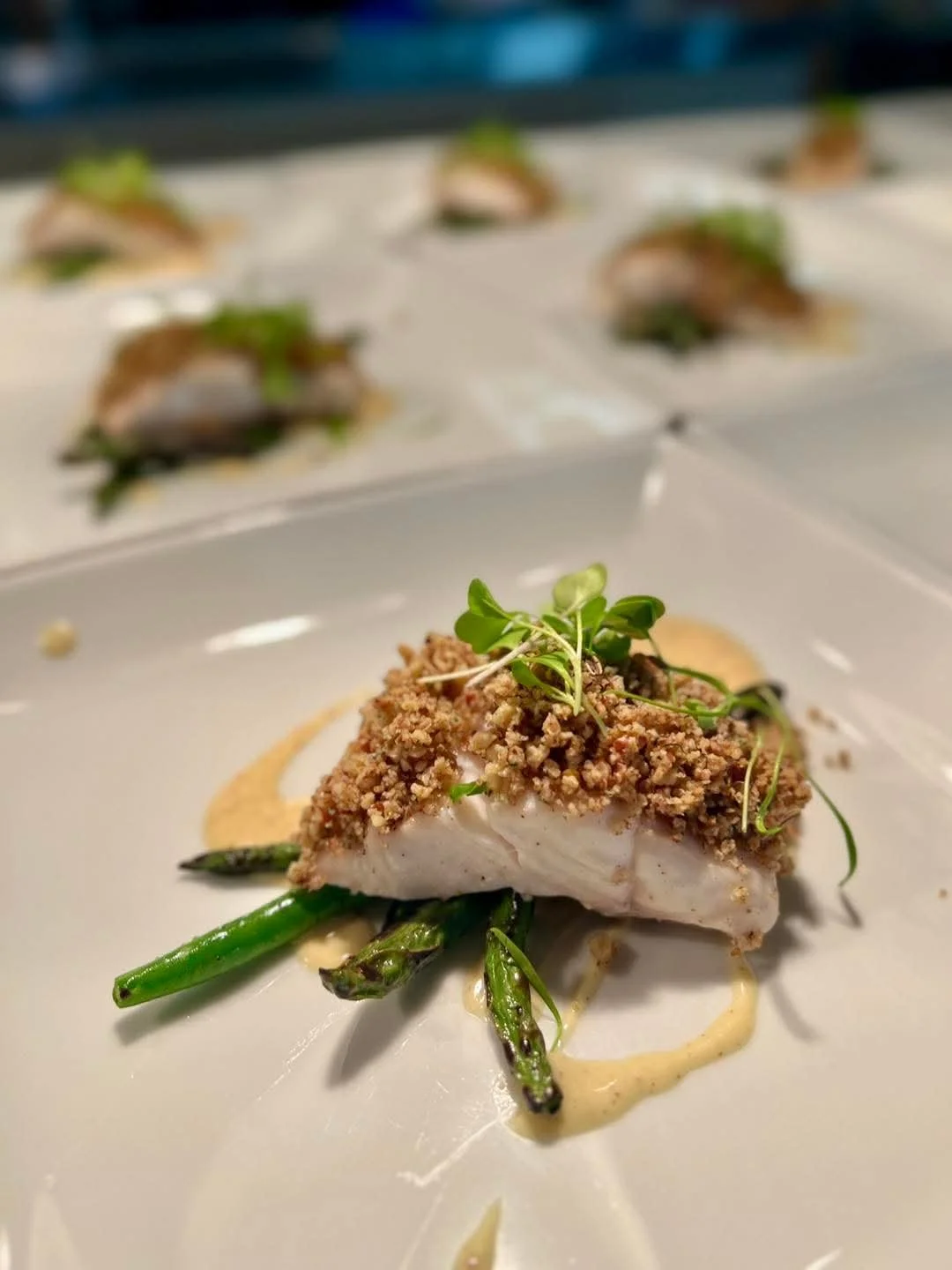 Gourmet dish with fish topped with breadcrumbs, served on grilled asparagus with sauce and microgreens on a white plate.