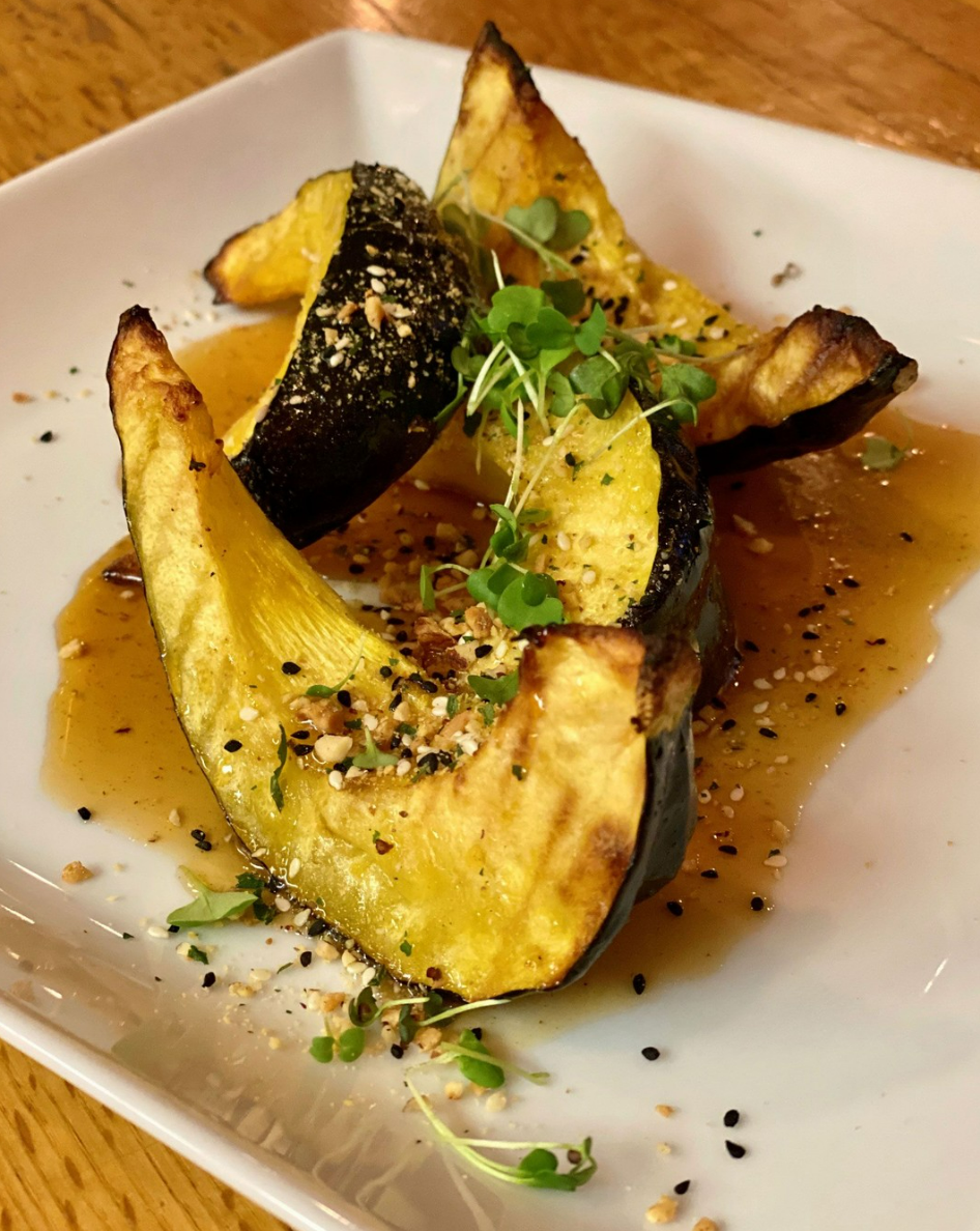 Roasted acorn squash slices on a white plate, garnished with green microgreens, sesame seeds, and a glaze sauce.