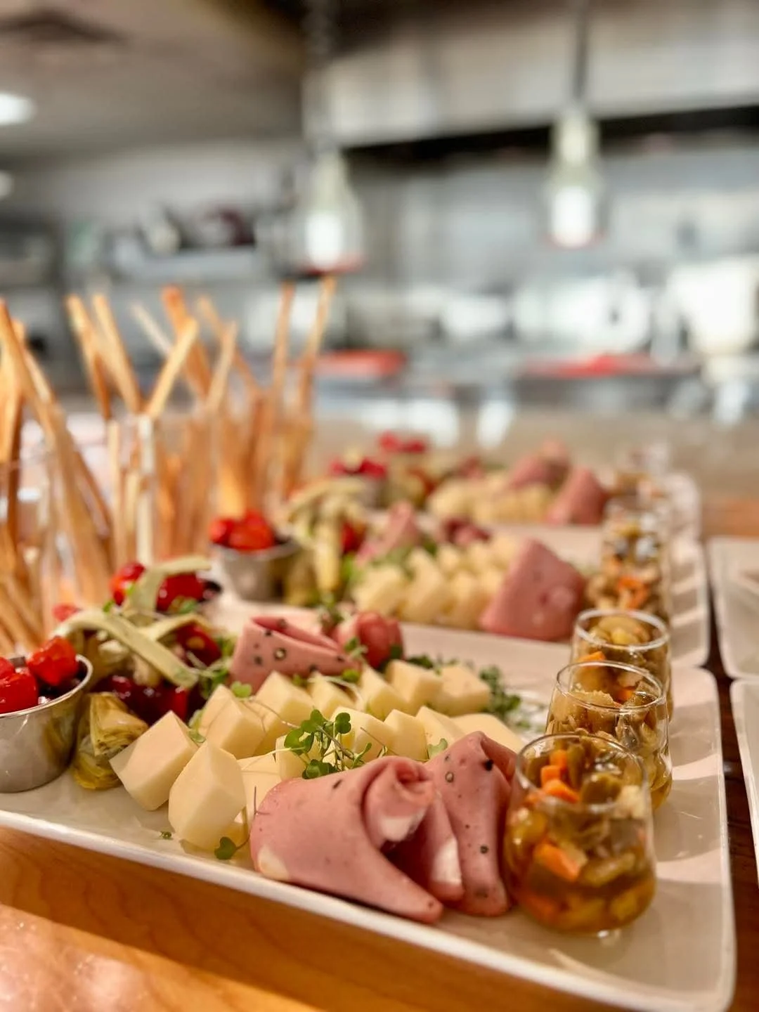 Close-up of a charcuterie board with cured meats, cheese cubes, pickled vegetables, cherry tomatoes, and breadsticks arranged on a white platter in a kitchen setting.