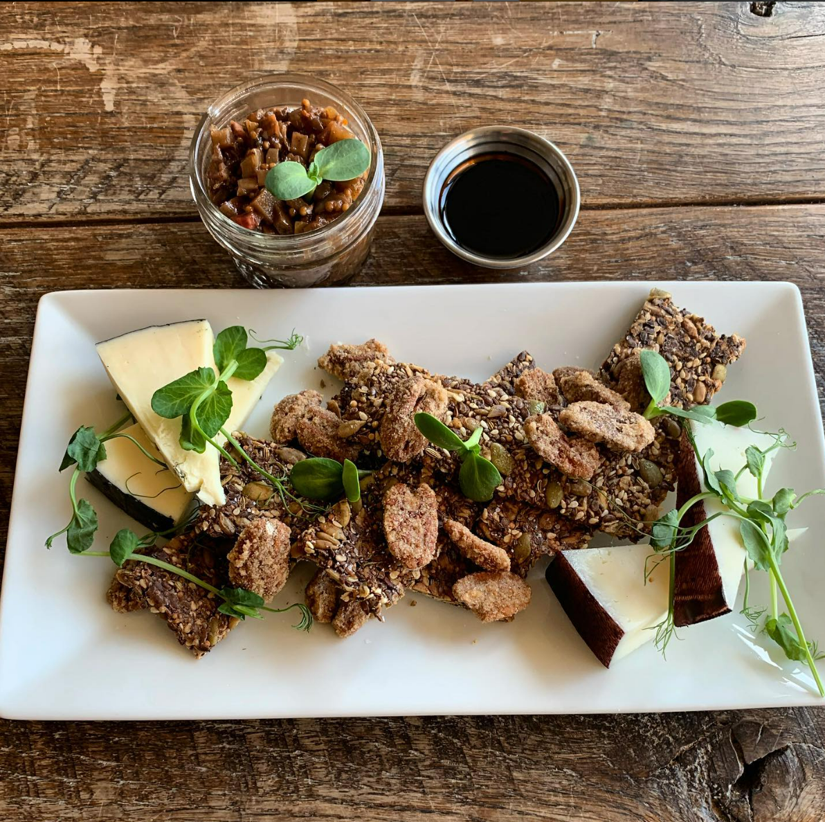 A platter with cheese wedges, seed crackers, caramelized nuts, microgreens, a small jar of chutney, and a bowl of dark sauce on a wooden table.