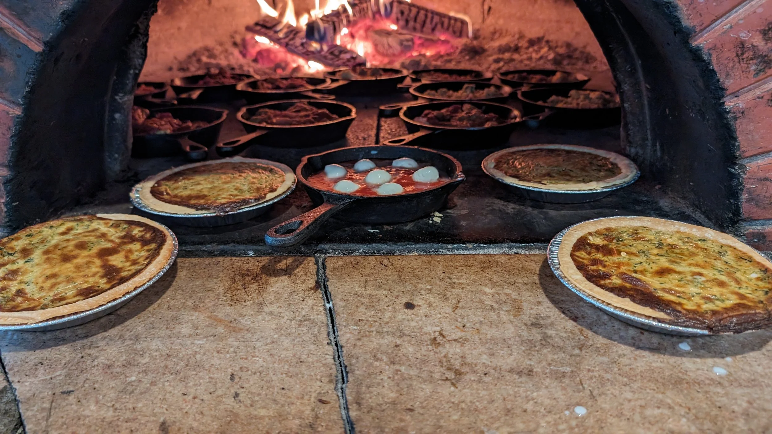 Wood-fired oven with quiches and a skillet of eggs cooking inside.