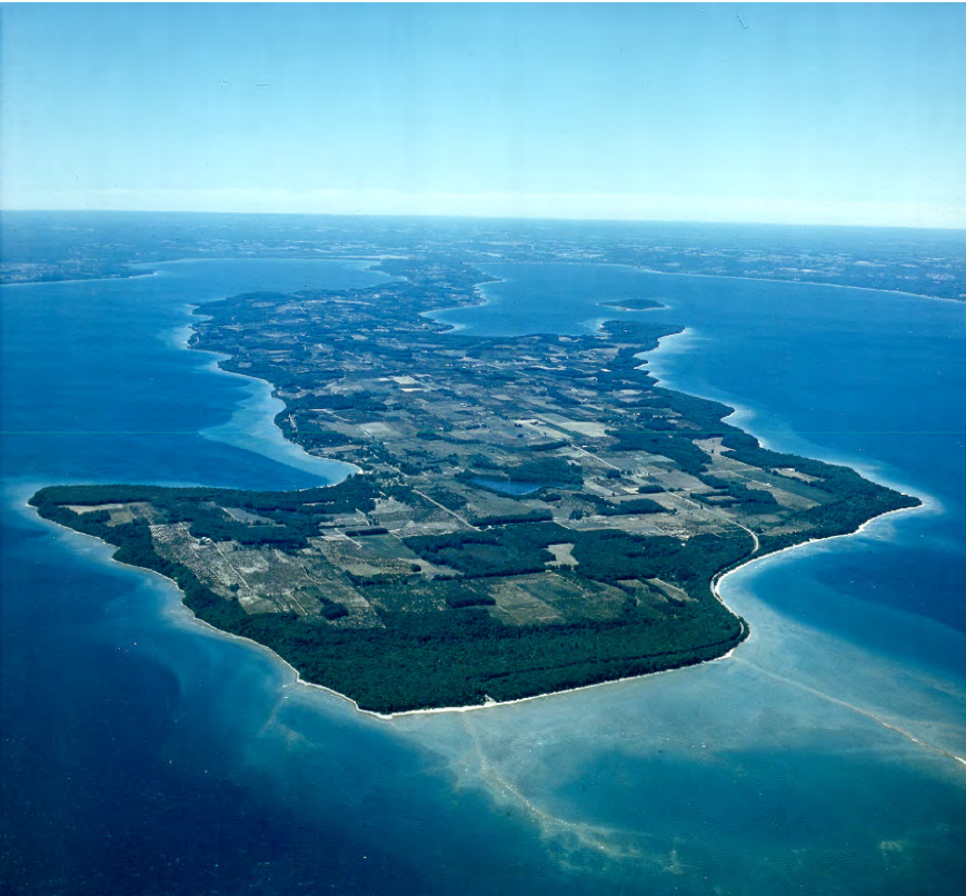 Aerial view of a large, narrow island with dense green forests and patches of open fields, surrounded by clear blue ocean waters.