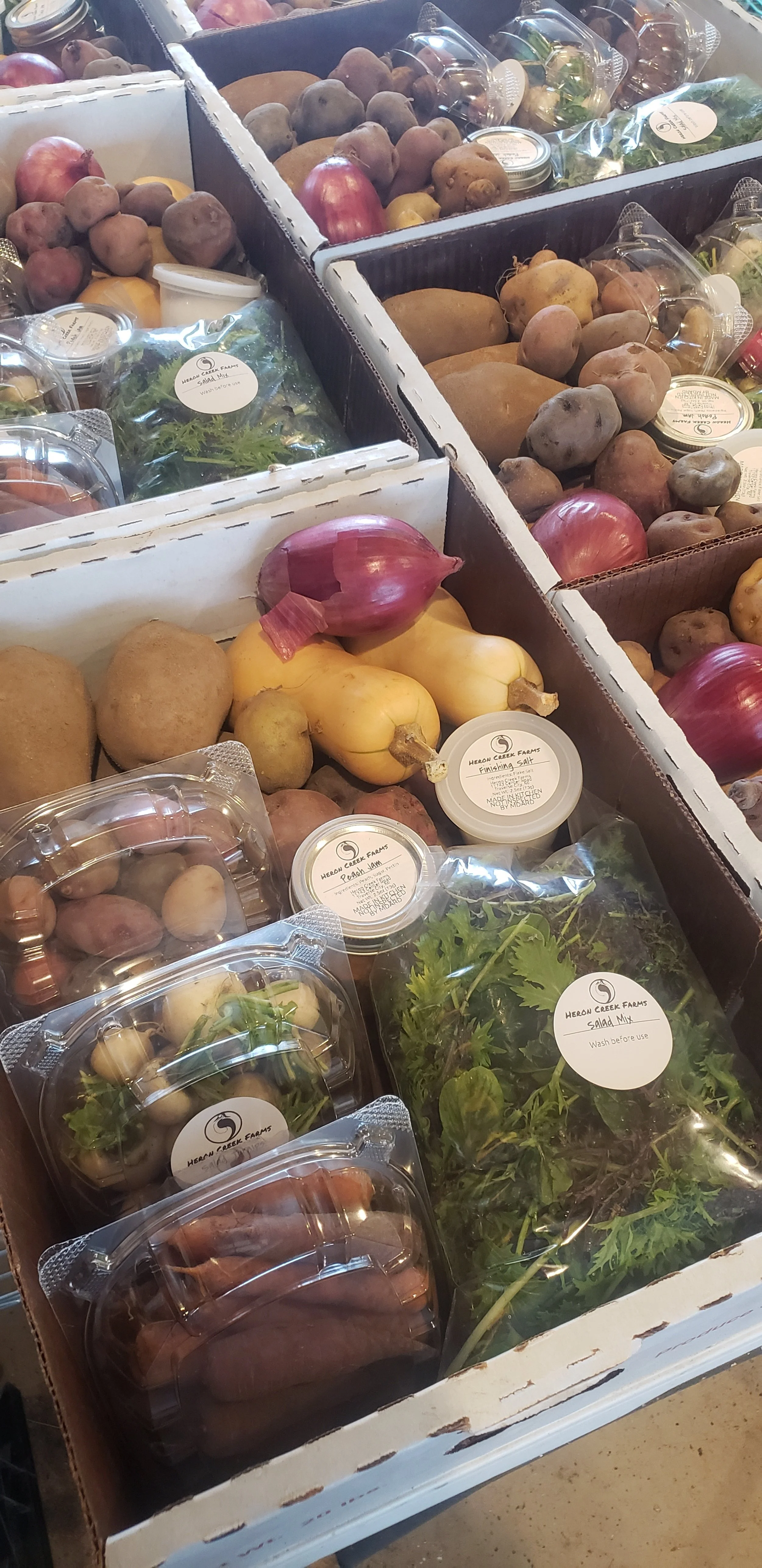Assorted fresh vegetables in boxes, including potatoes, onions, squash, greens, and packaged produce with Heron Creek Farms labels.