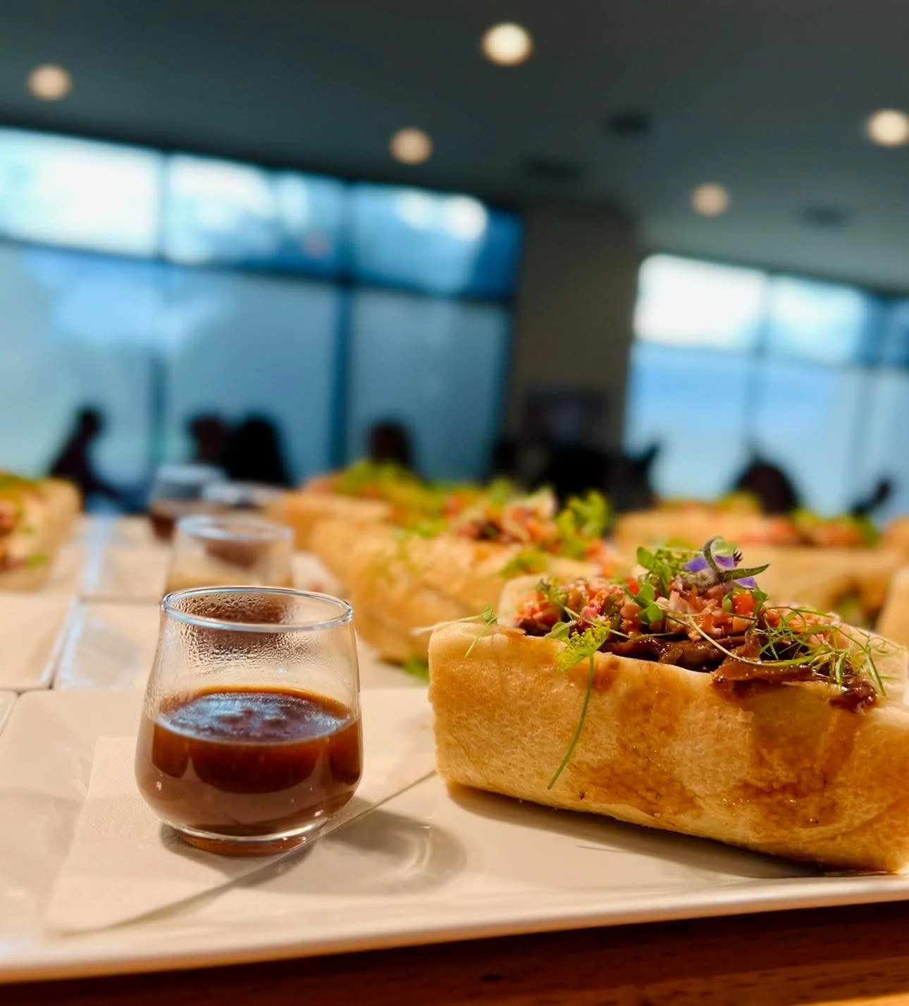 A close-up of a sandwich filled with shredded meat and garnished with herbs on a white plate with a small glass of dark sauce. Blurred people and windows in the background.