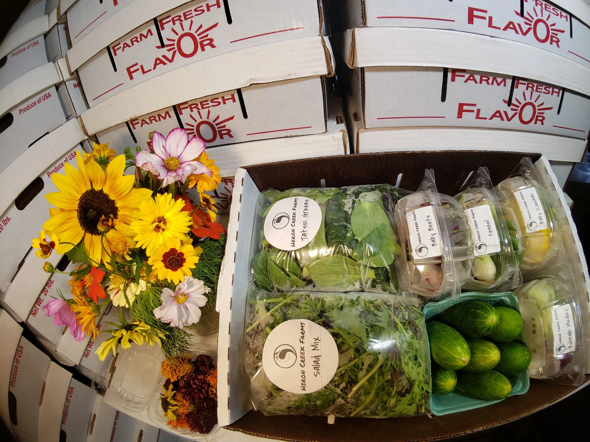 Box of fresh produce and flowers, including leafy greens, squash, beets, cucumbers, and a colorful bouquet, with stacked boxes labeled 'Farm Fresh Flavor' in the background.