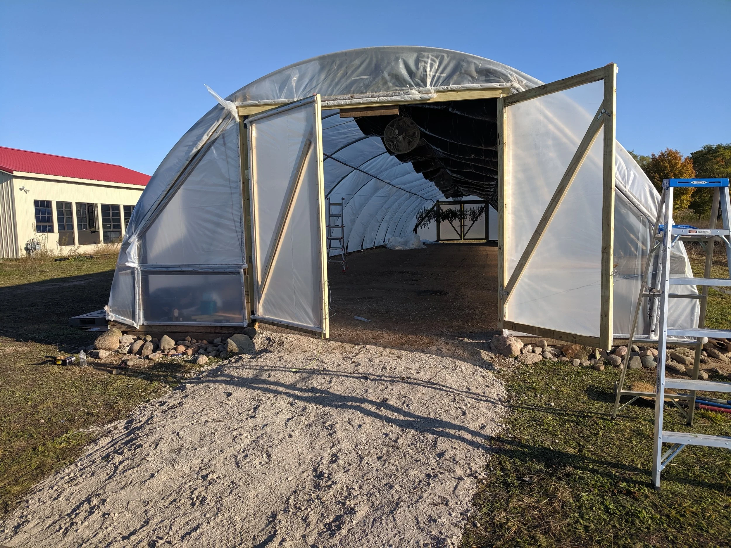Open-ended hoop house structure with plastic covering and a metal ladder outside, surrounded by rocks and grass.