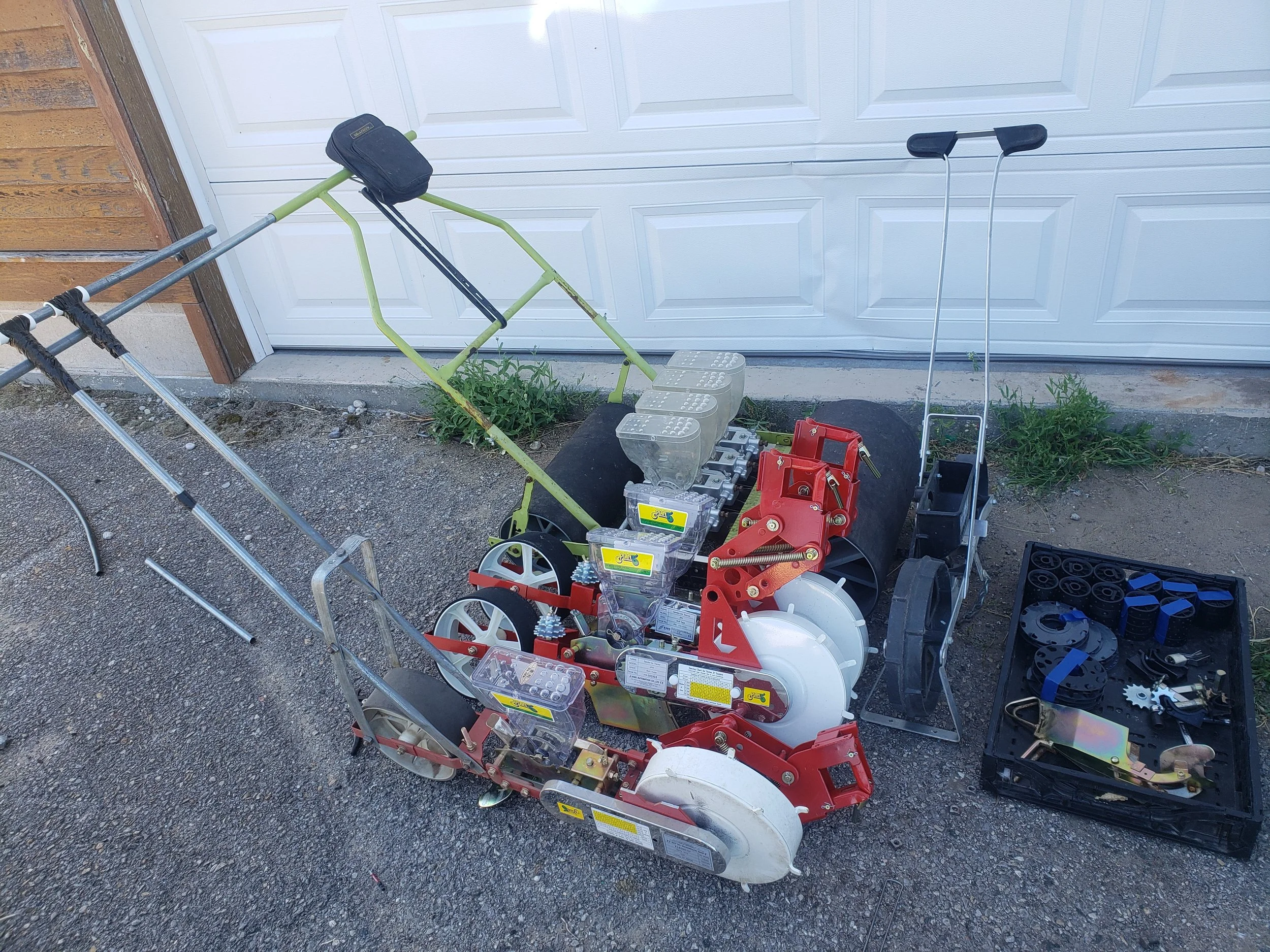 Garden tools and equipment including a manual push seeder, trays with seed plates and accessories, placed on a driveway next to a garage door.