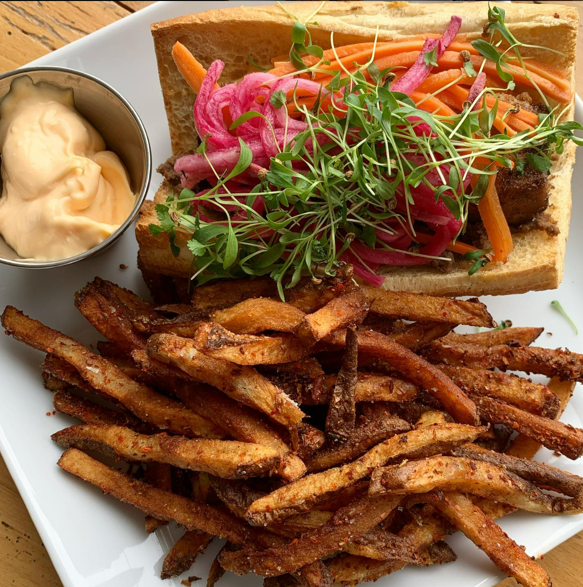 Plate with a sandwich, French fries, and dipping sauce. The sandwich features pickled onions, carrots, greens, on a baguette. Fries are seasoned and crispy. A small cup of dipping sauce accompanies the meal.