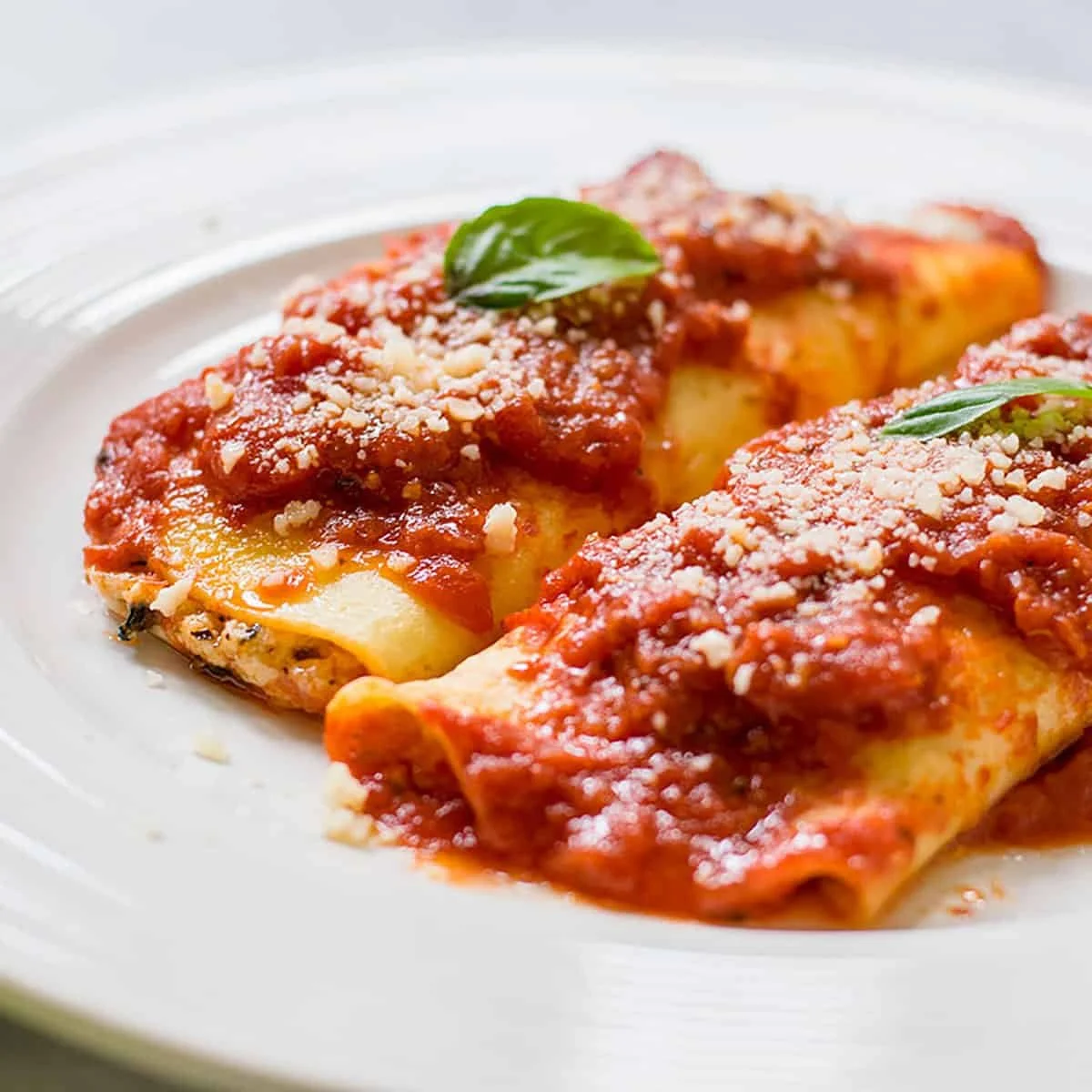 Two cannelloni pasta rolls topped with marinara sauce, grated cheese, and basil on a white plate.