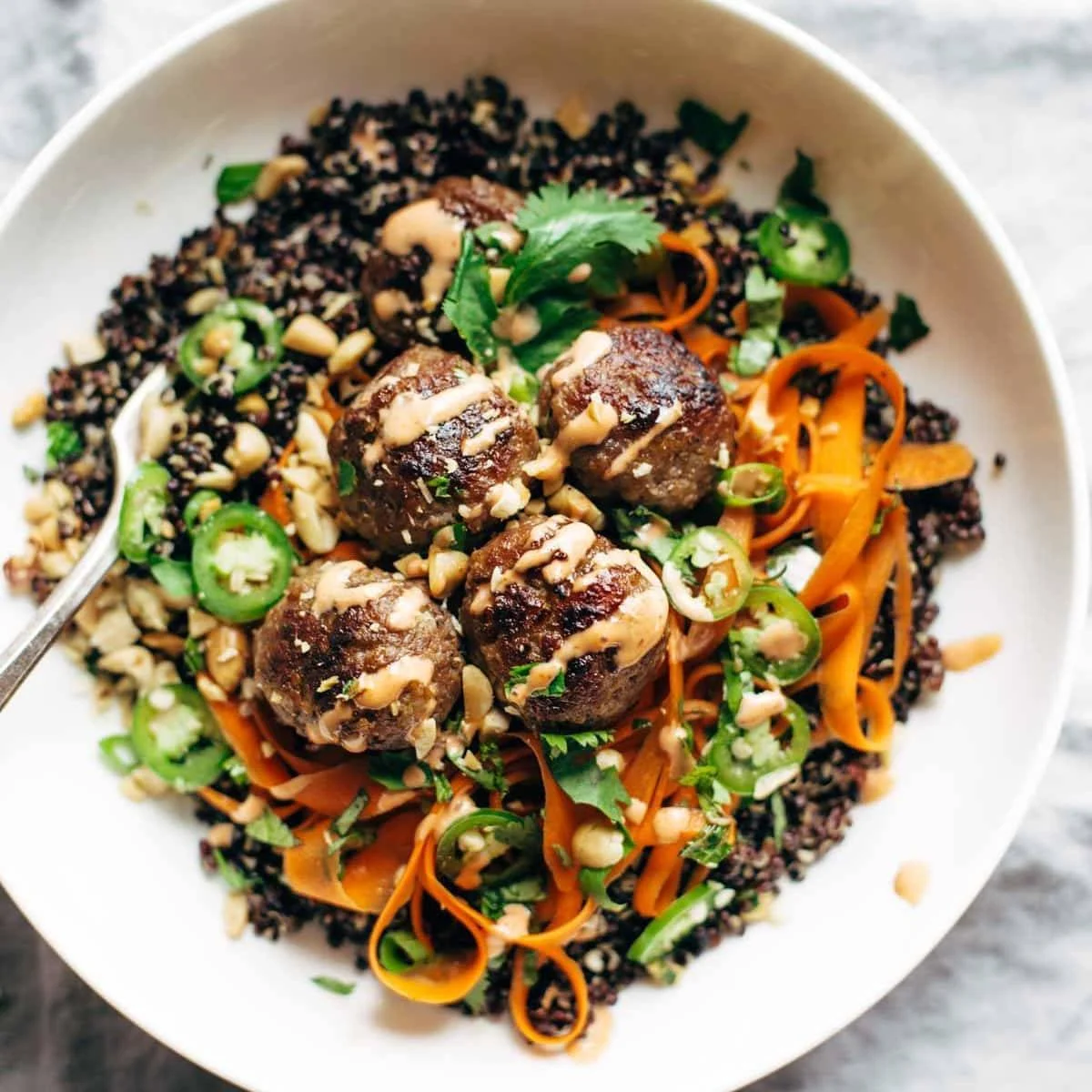 A bowl of quinoa topped with meatballs, julienned carrots, sliced jalapeños, fresh cilantro, and a creamy sauce, garnished with chopped peanuts.