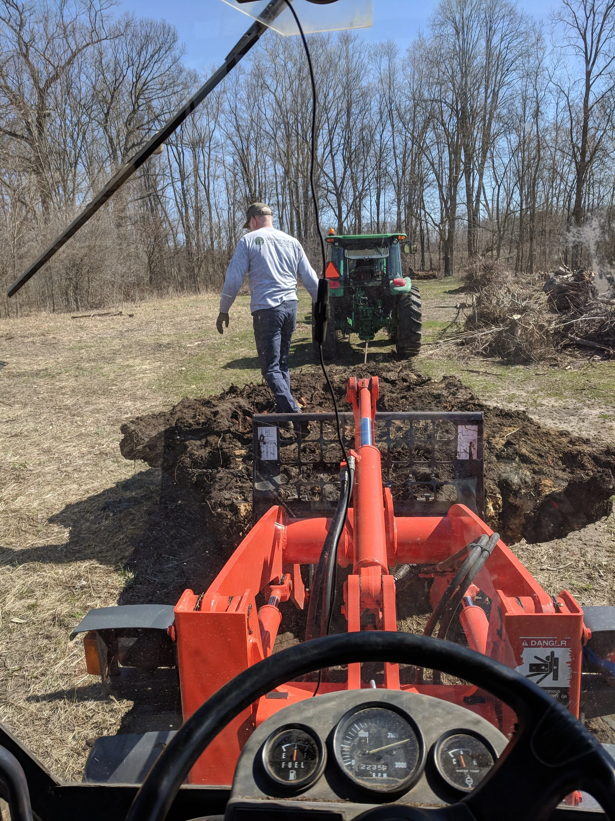 View from inside a tractor cab showing another person walking towards a green tractor on a rural field, with tree line in background, on a clear day.