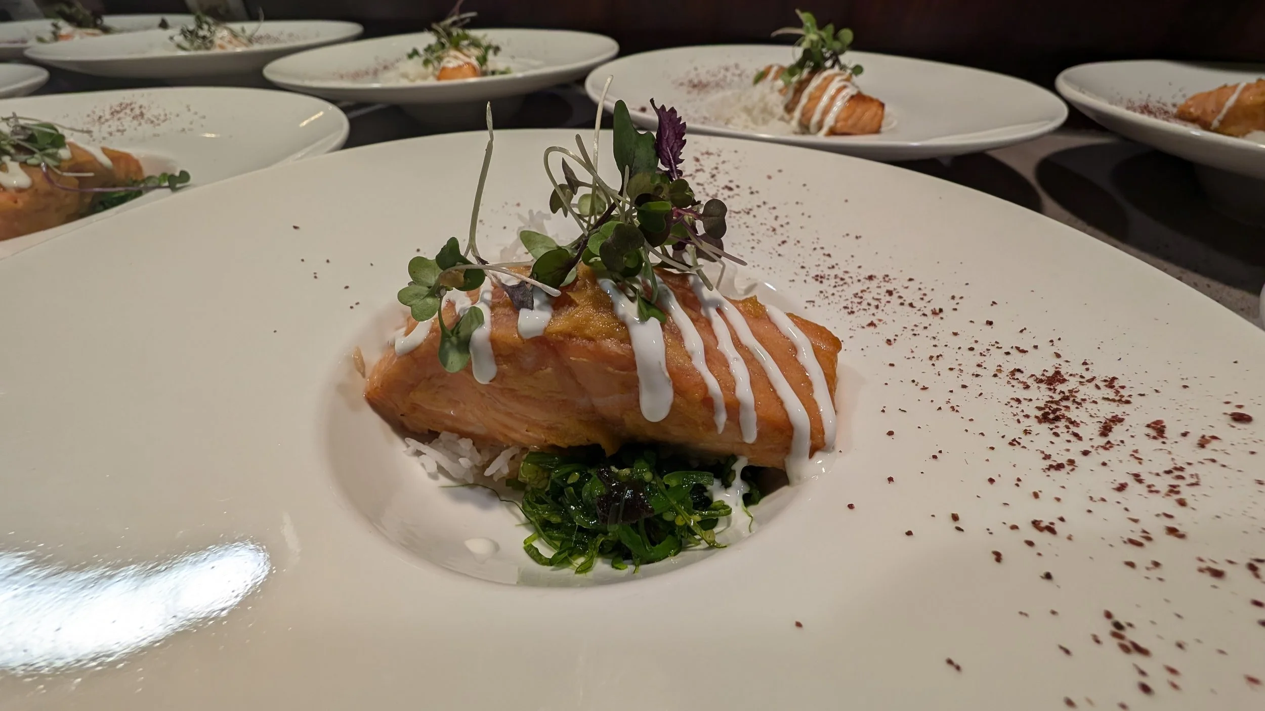 Plated dish with cooked salmon topped with a creamy drizzle and microgreens, served on a bed of rice and vegetables, garnished with seasoning, arranged with similar plates in the background.