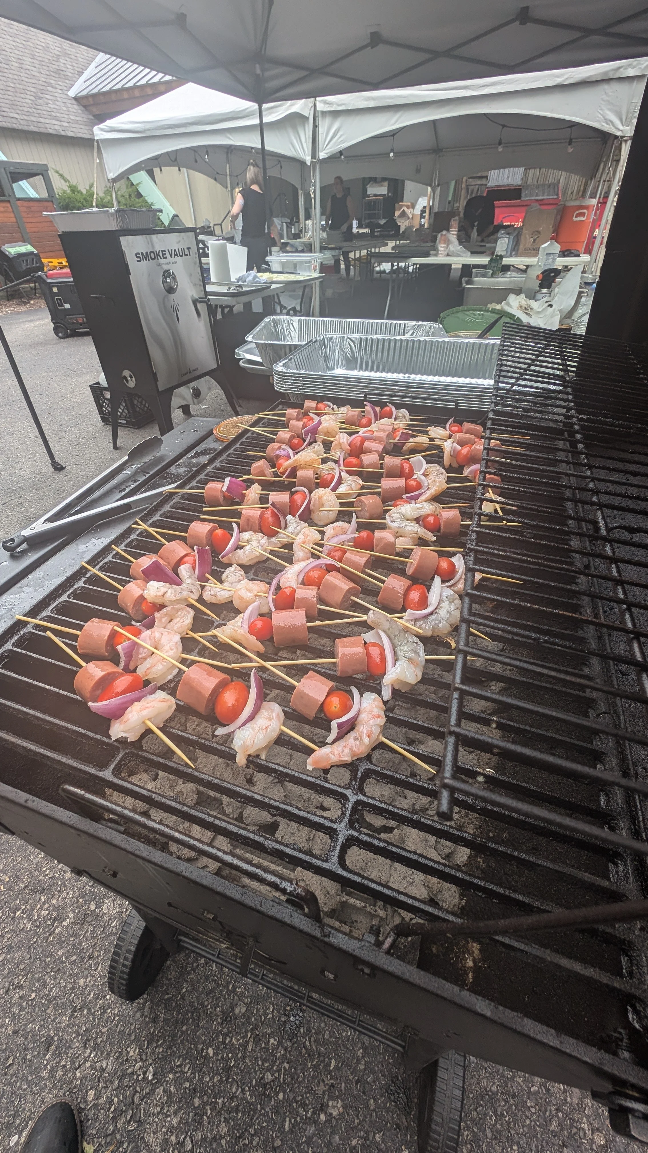 Outdoor grill with skewers of shrimp, sausage, cherry tomatoes, and onions under a canopy tent with people preparing food in the background.
