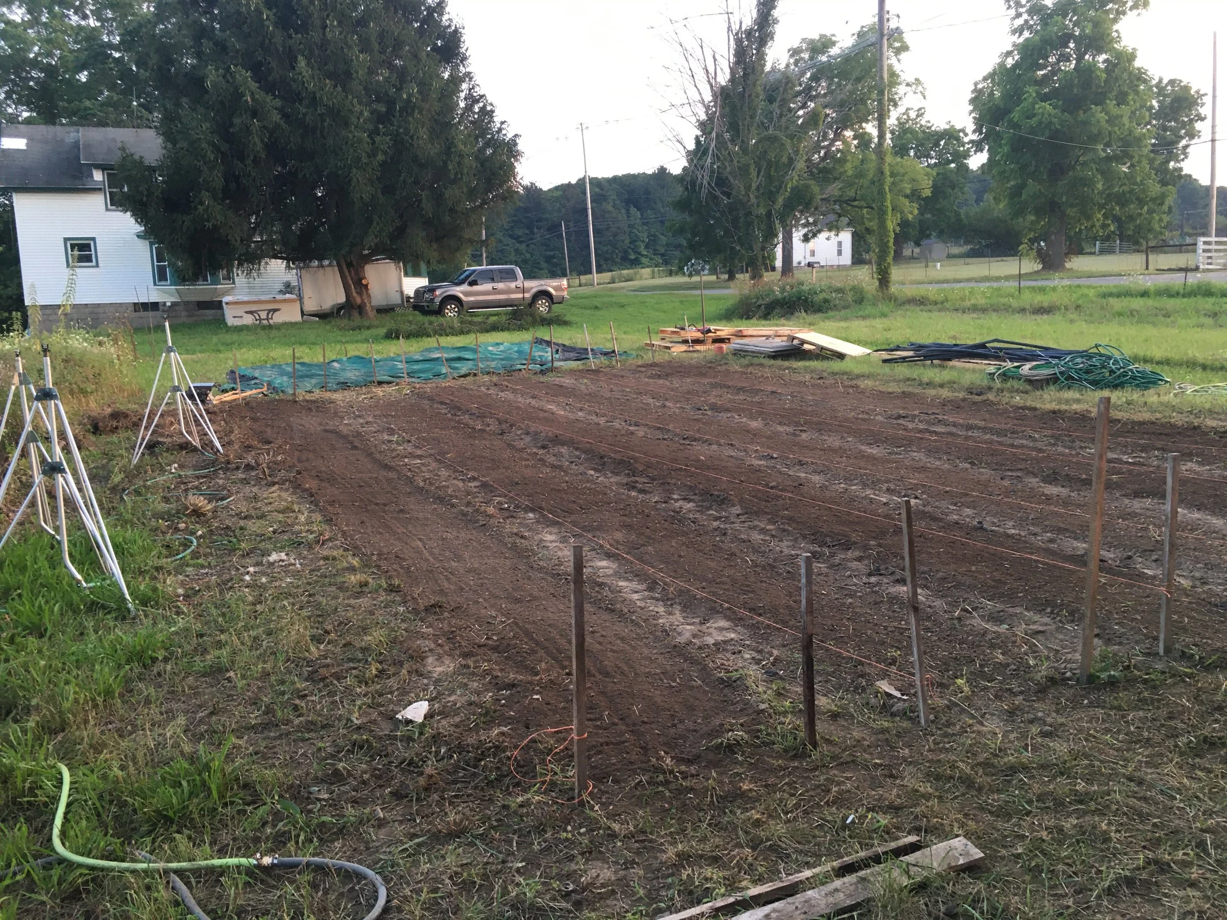 Freshly tilled garden plot on a farm with a house, truck, and large tree in the background. Hoses and equipment are scattered around the area.
