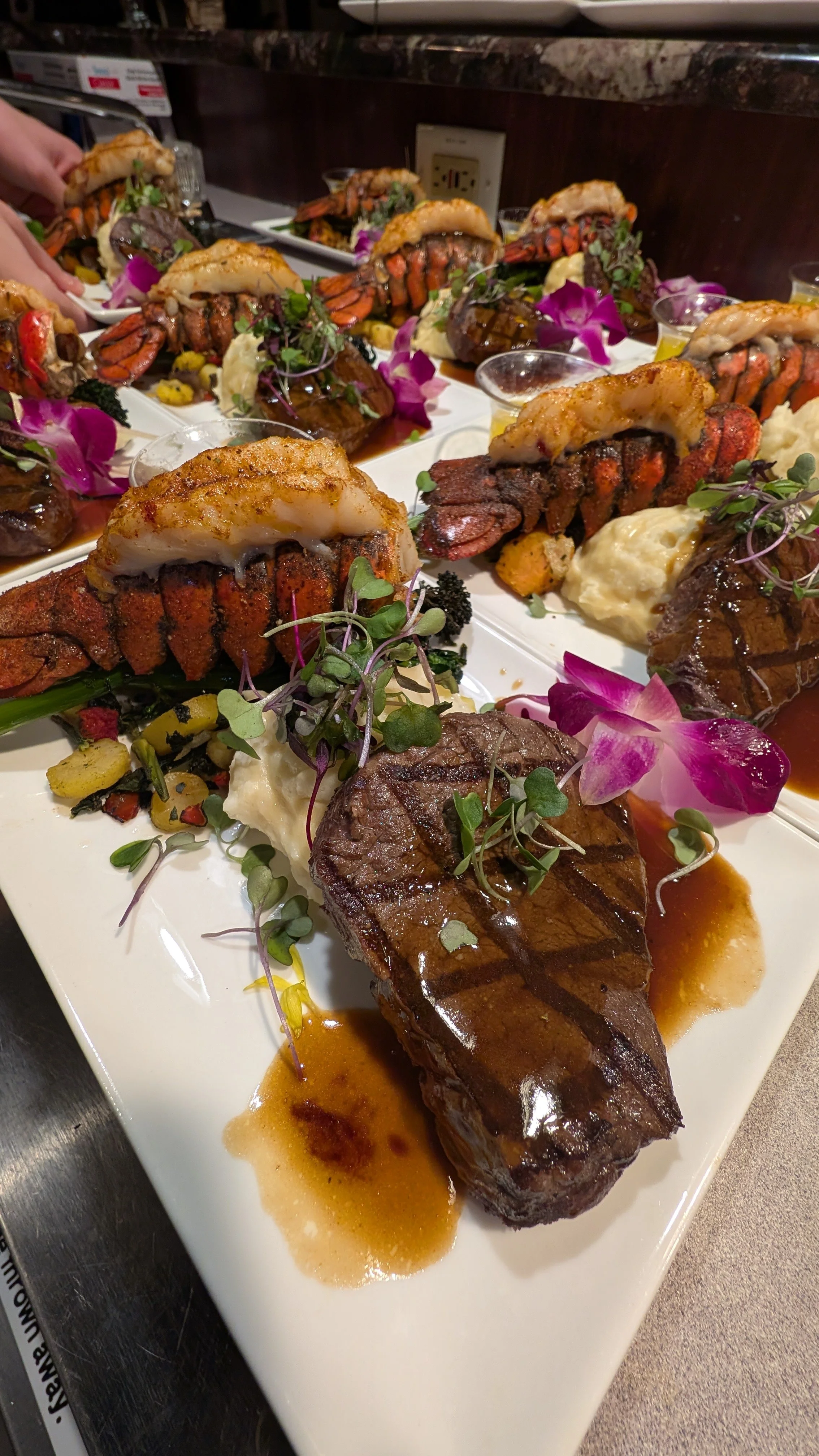 Plate of surf and turf featuring grilled lobster tails, steak, microgreens, mashed potatoes, and edible flowers.