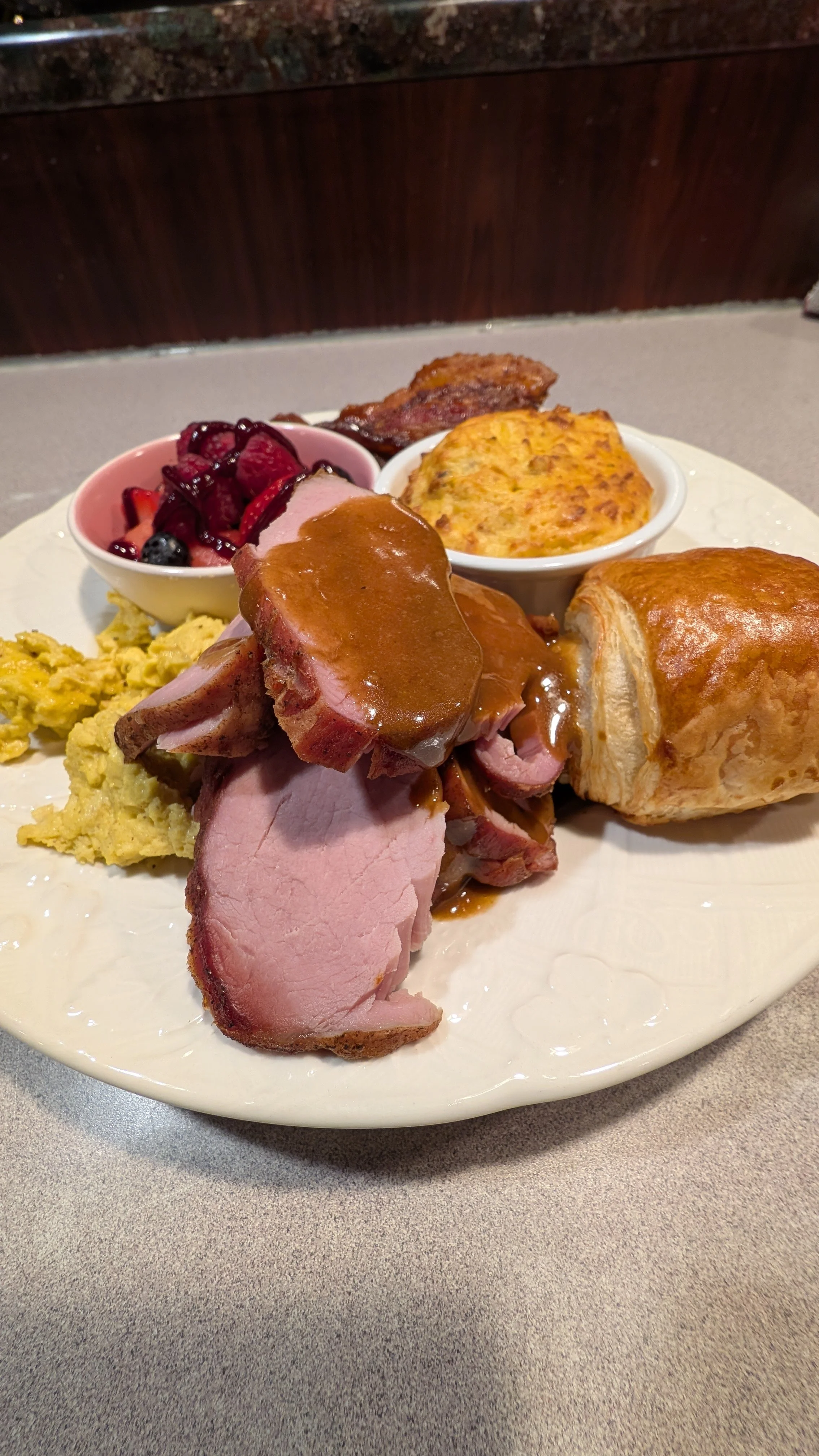 A plate of sliced roasted meat with gravy, scrambled eggs, a pastry, a small bowl of fruit, and a baked savory dish.