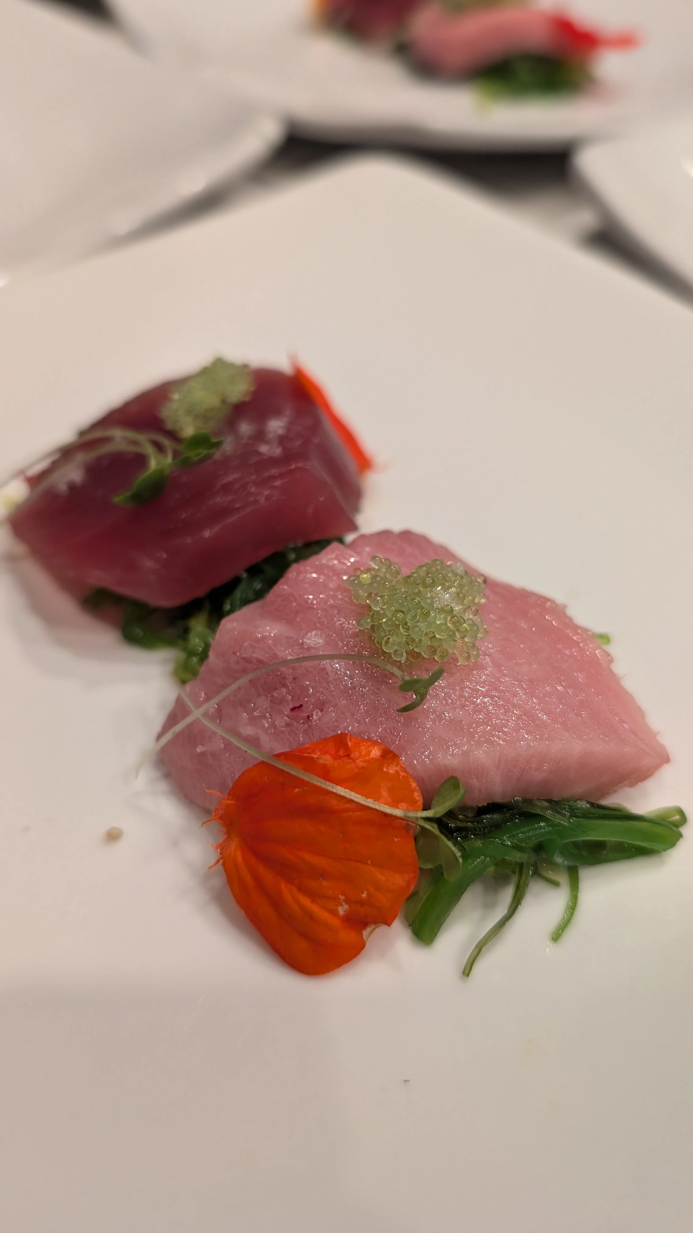 Sashimi dish with tuna, edible flower, and garnish on white plate.