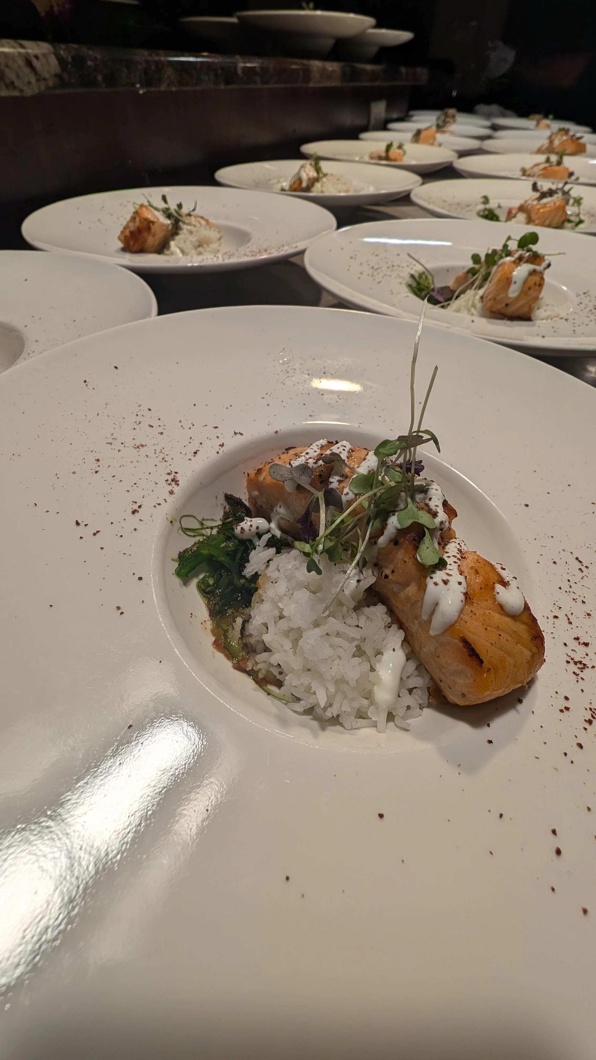 Plated salmon with rice, greens, microgreens, and sauce in a restaurant setting.