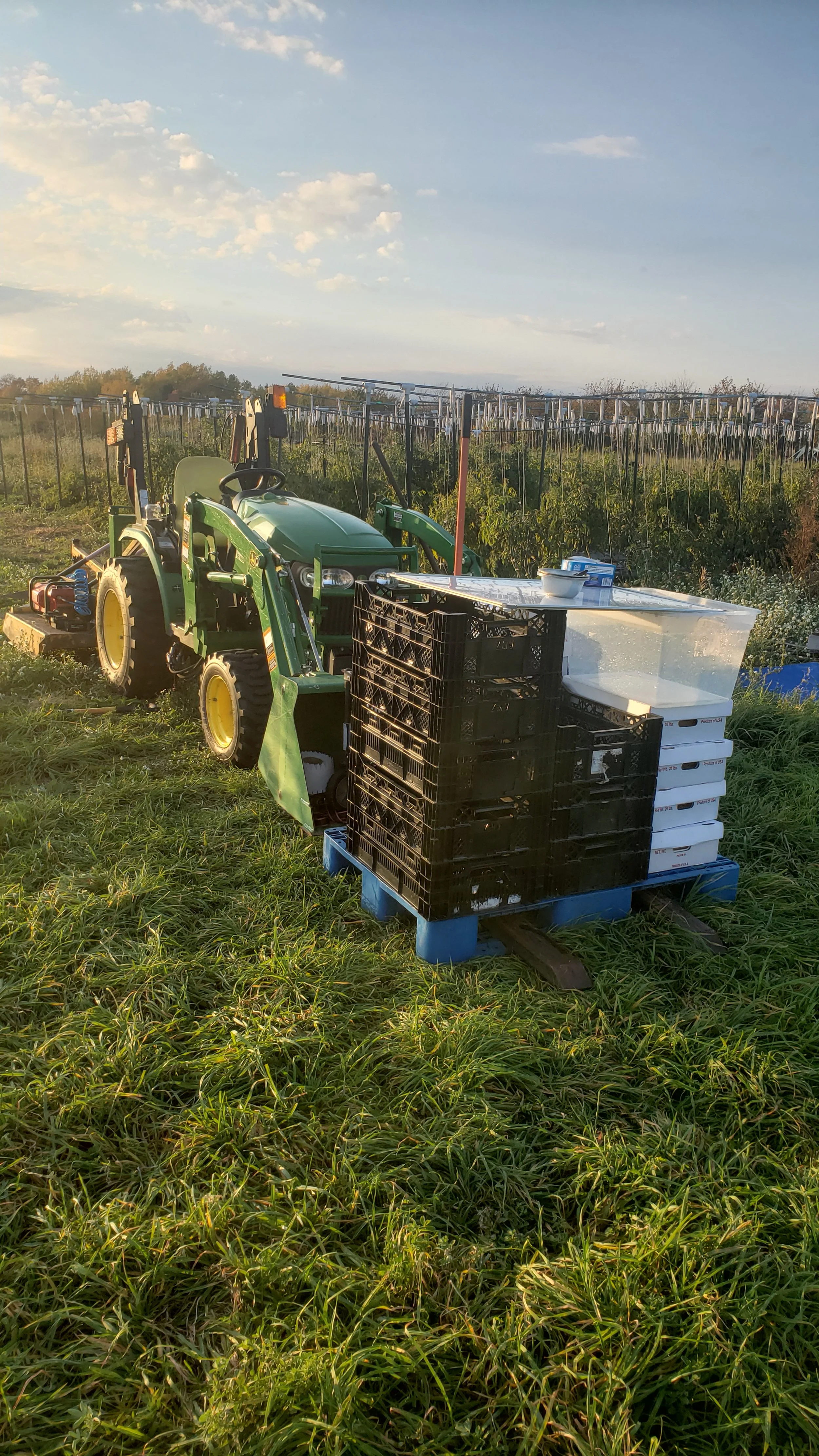 Green tractor with attachments parked on a grassy field next to stacked plastic boxes and crates on a blue pallet, with solar panels visible in the background under a clear sky.