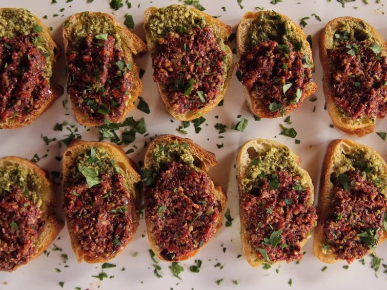 Slices of toasted bread topped with pesto and a minced olive mixture, garnished with chopped parsley on a light surface.