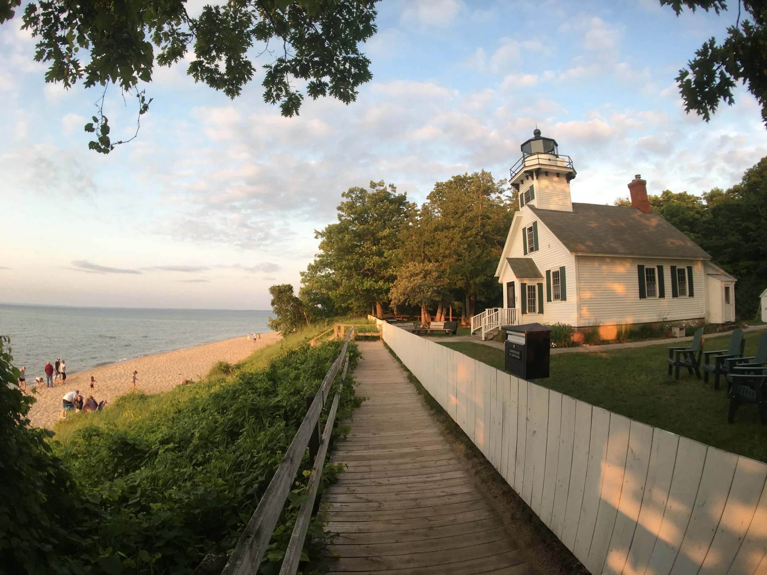 A scenic view of a lighthouse near a beach, surrounded by trees and foliage. People are walking on the sandy shore and relaxing in the area. A wooden walkway leads towards the lighthouse, which has a traditional design with a white exterior and black