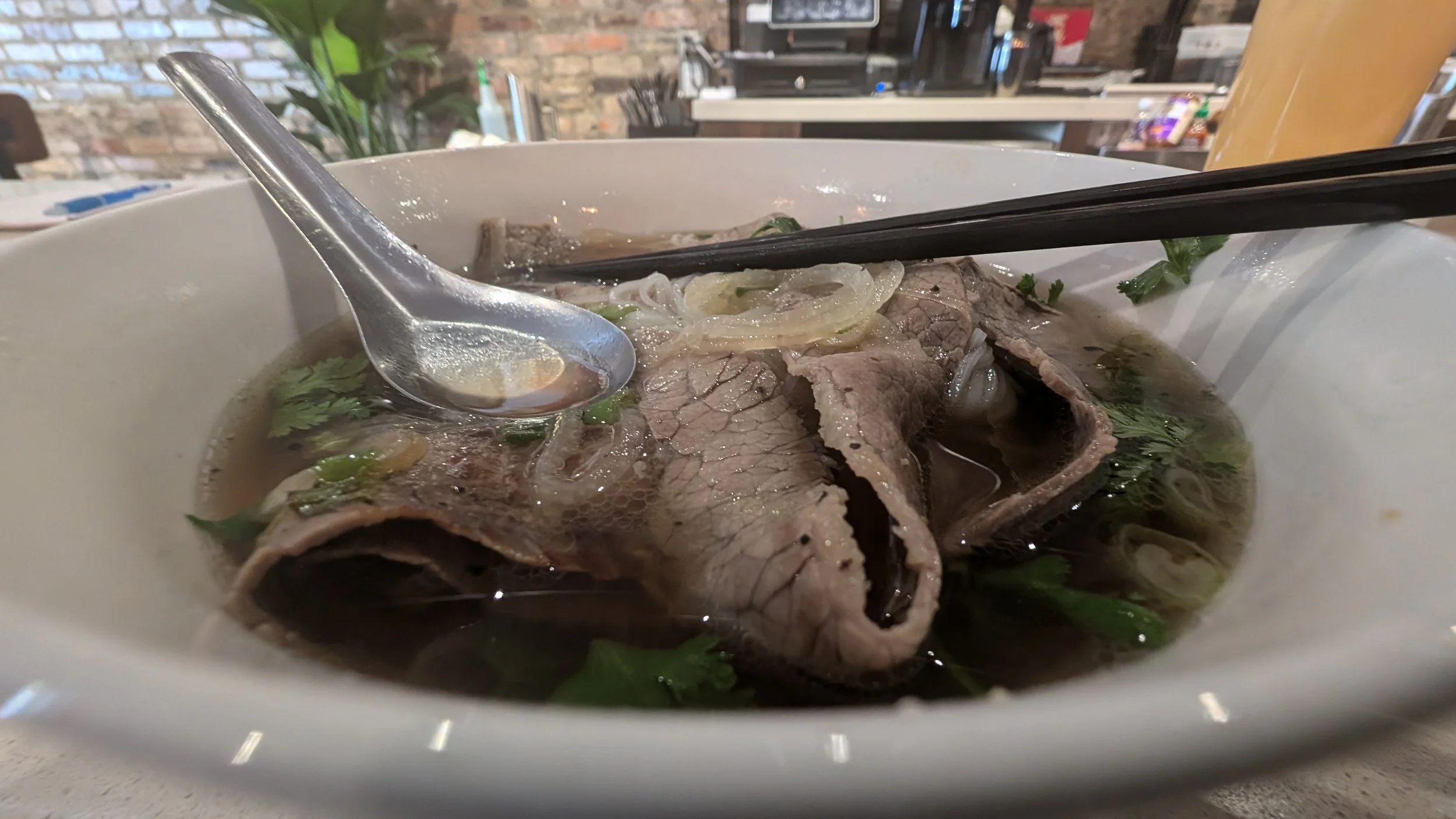 A bowl of Vietnamese pho with beef, onions, cilantro, and a metal soup spoon, with chopsticks resting on the bowl.