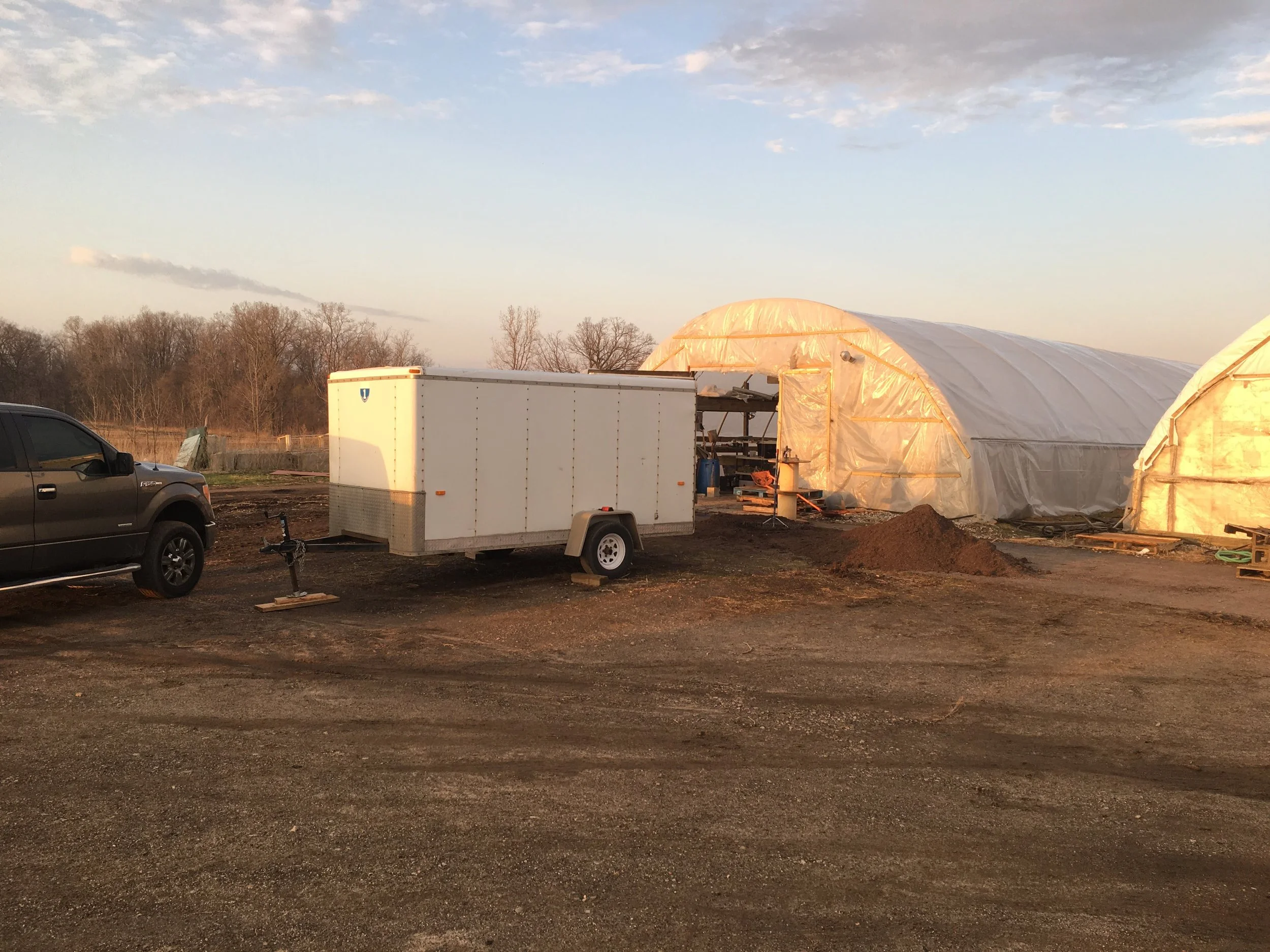 Outdoor scene with a white utility trailer, a parked dark-colored truck, and two large white greenhouse structures on a dirt field.