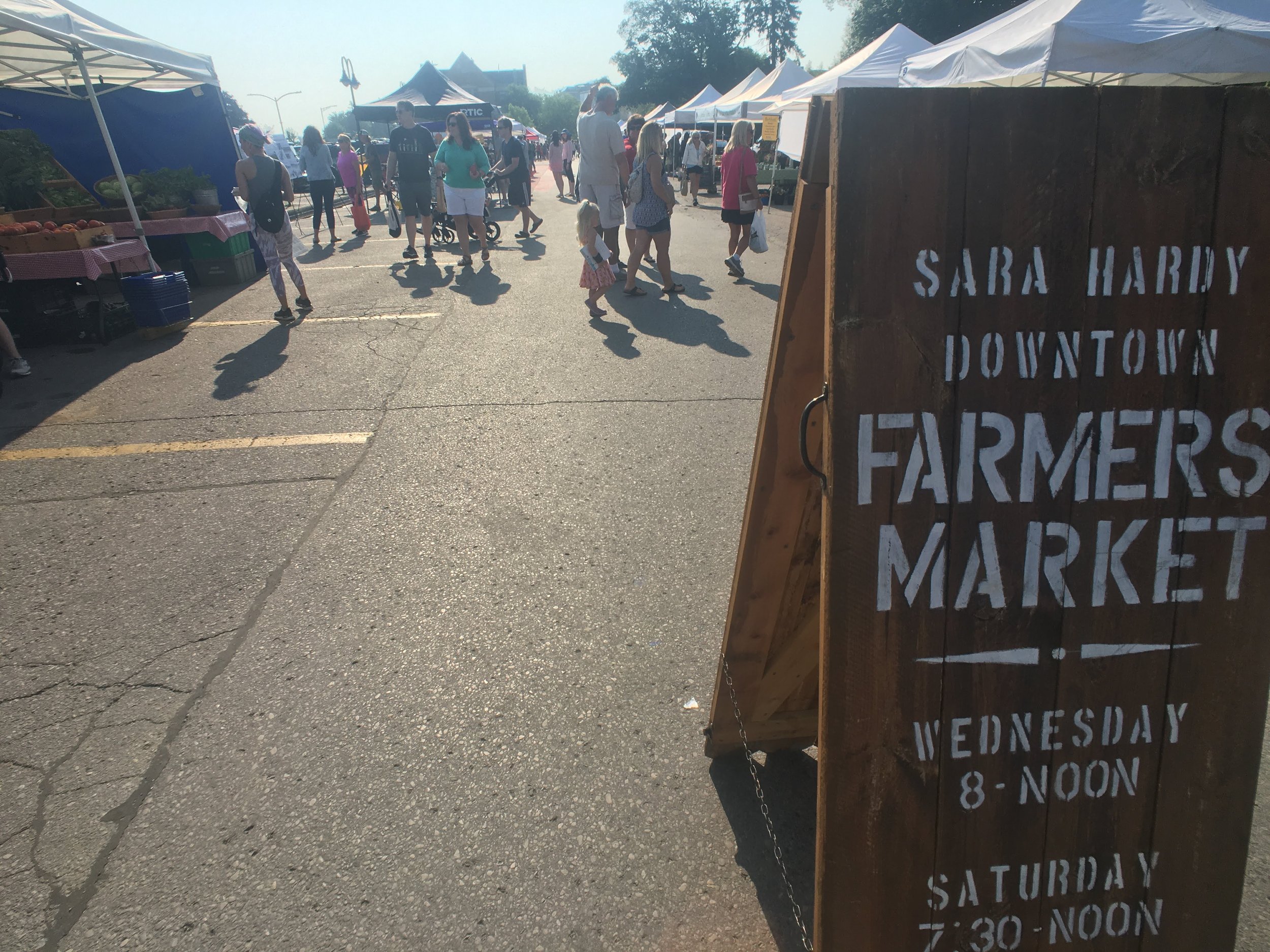 People walking at an outdoor farmers market with a wooden sign saying "Sara Hardy Downtown Farmers Market." Various vendor tents and tables are visible.