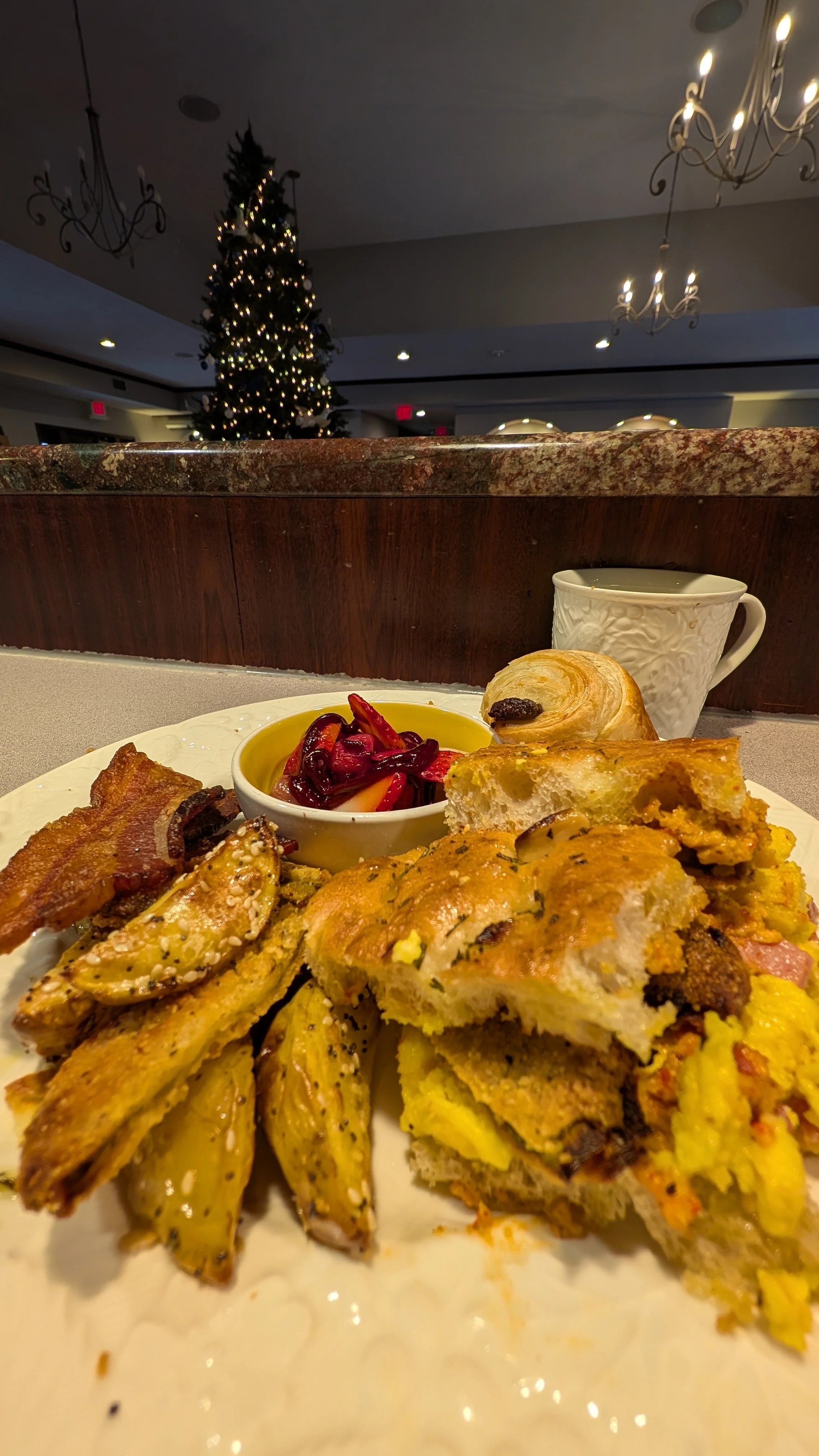 A plate of breakfast food with egg sandwich, bacon, seasoned potato wedges, and a bowl of red peppers. In the background, a cup and a Christmas tree.