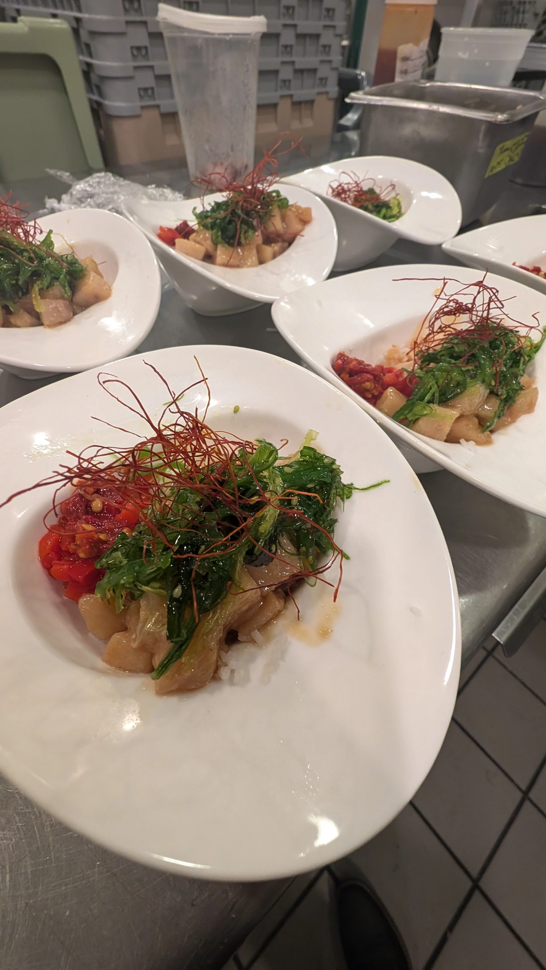 Plates of gourmet dishes with leafy greens, diced fish, red sauces, and garnished with thin red strands in a kitchen setting.