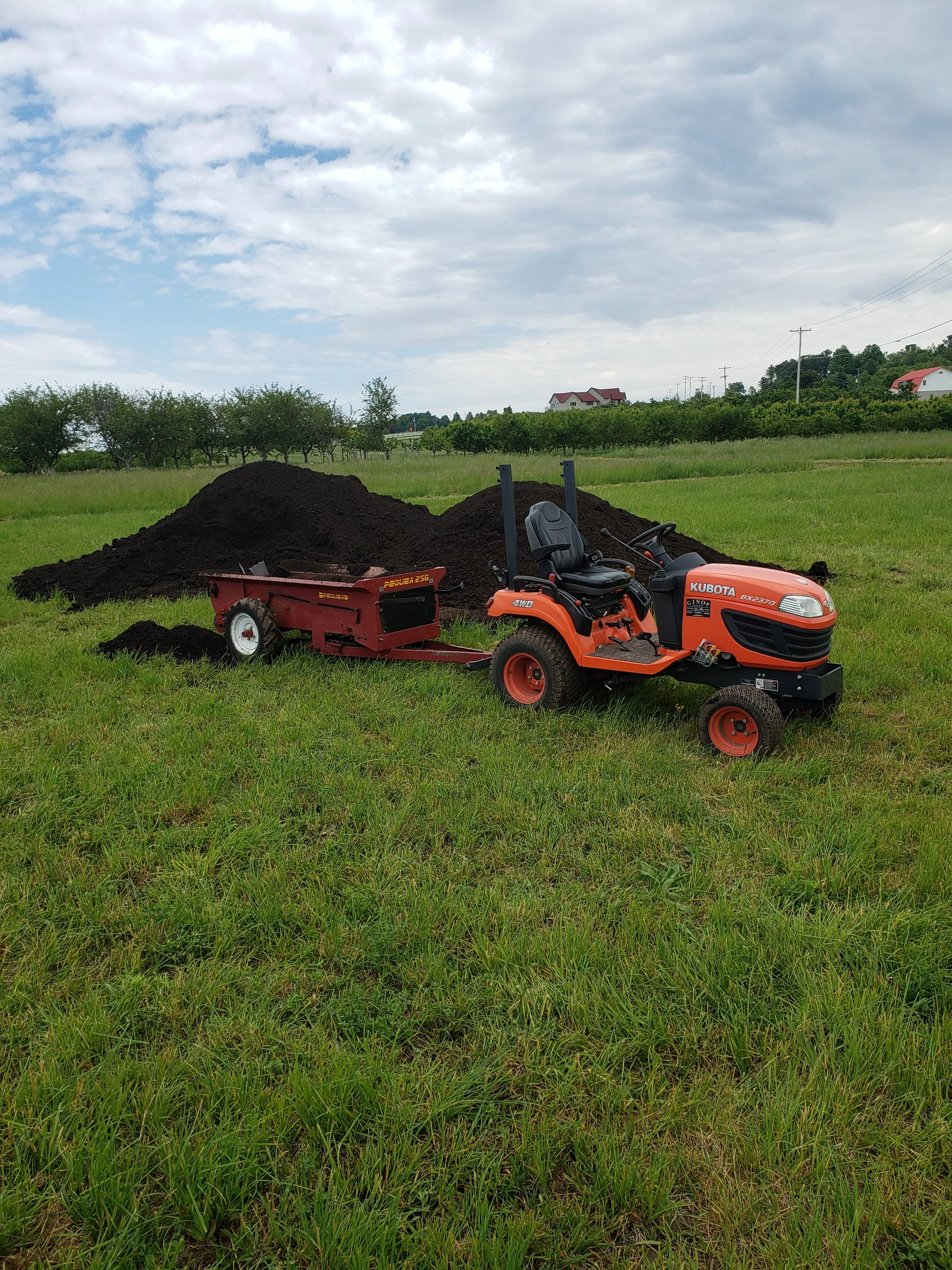 Orange tractor with a trailer attachment on a grassy field beside a large pile of dark soil, under a partly cloudy sky.