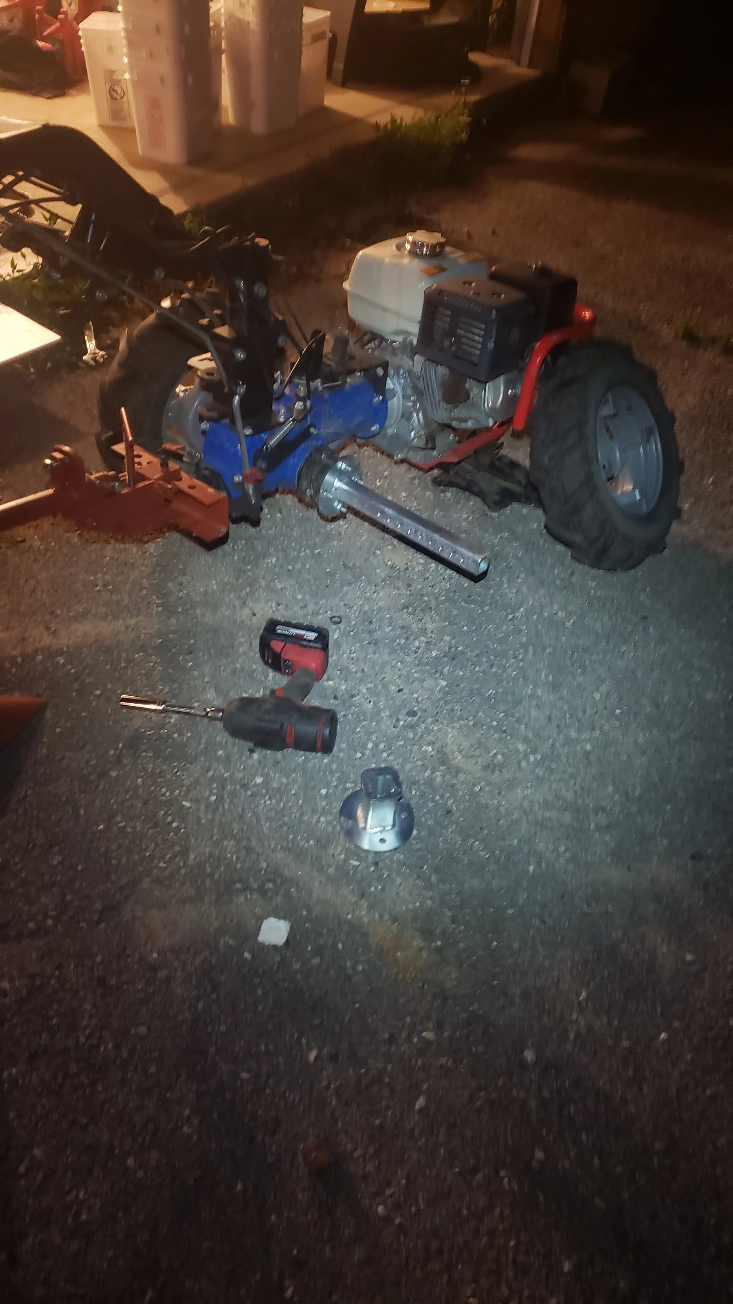 Nighttime scene showing a small engine machine with a tiller or cultivator attachment, power tools, and plastic containers in the background.