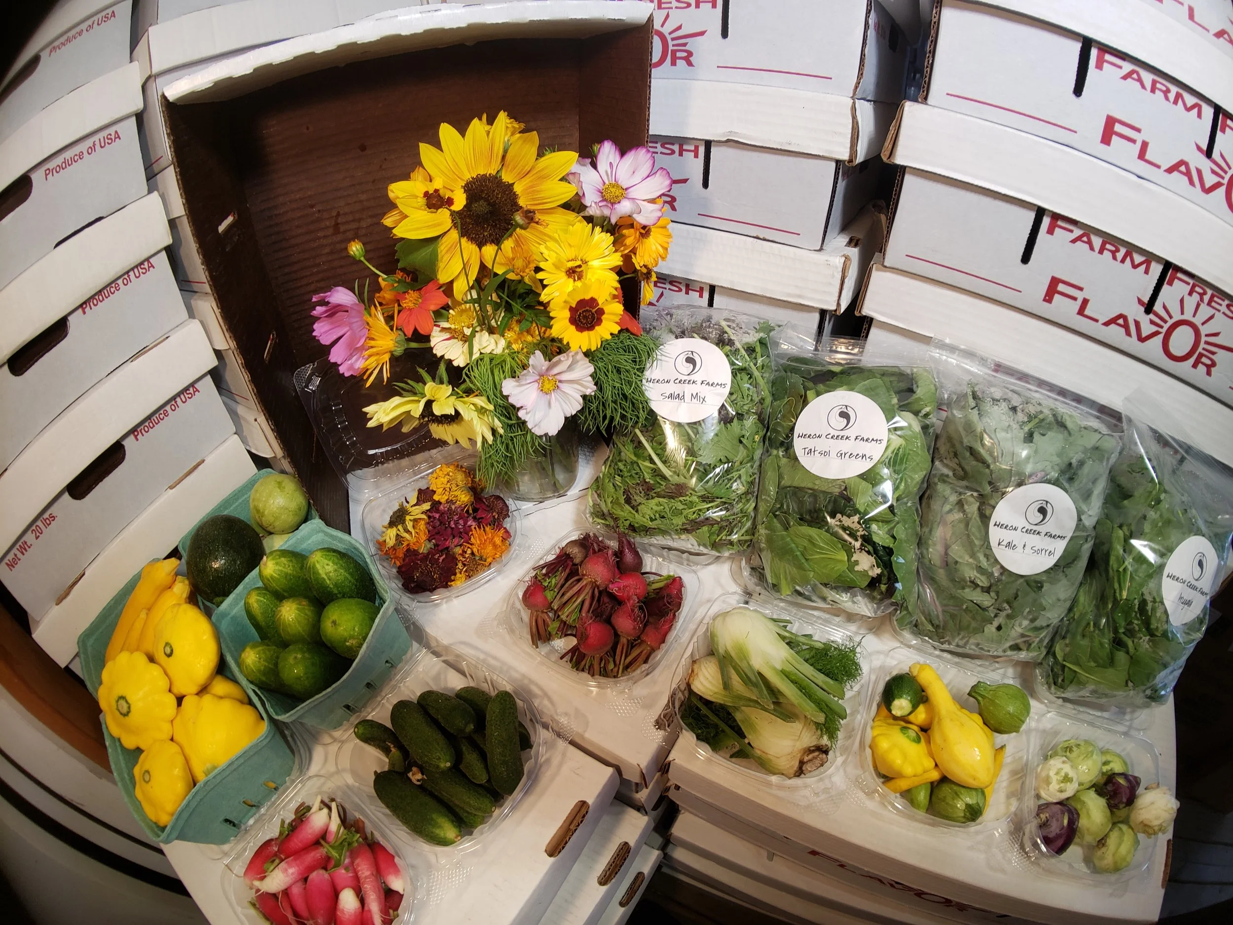 Assorted fresh vegetables including squash, cucumbers, radishes, greens, and a bouquet of flowers on display at a produce market. Packaged leafy greens from Heeren Creek Farms are also shown, with boxes labeled "Produce of USA" in the background.