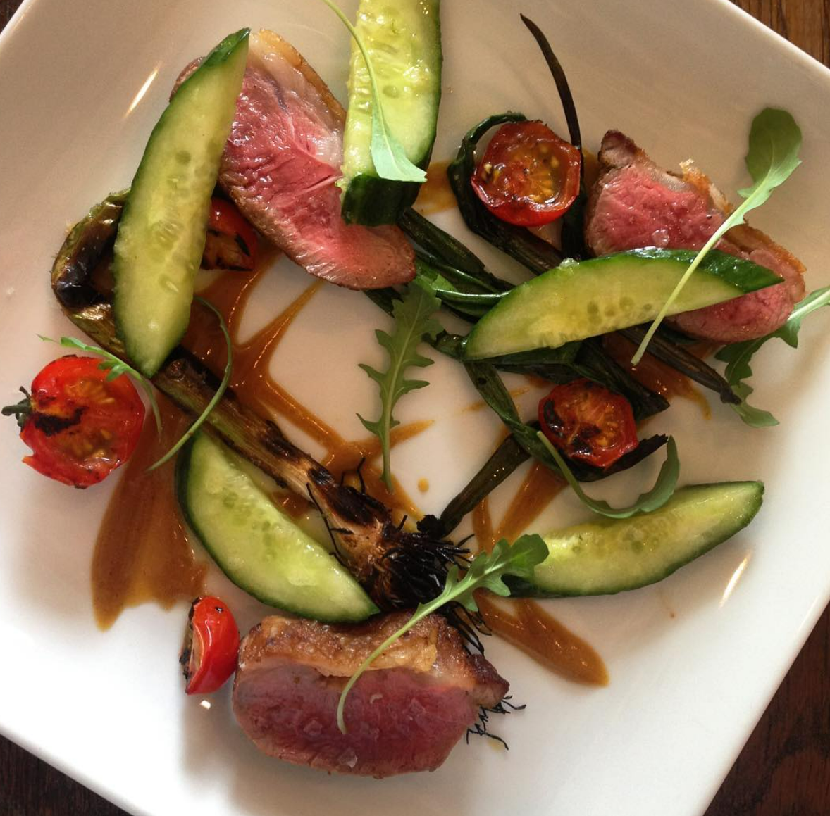 Gourmet dish with sliced medium-rare duck breast, cherry tomatoes, cucumber slices, grilled scallions, garnished with arugula on a white plate.