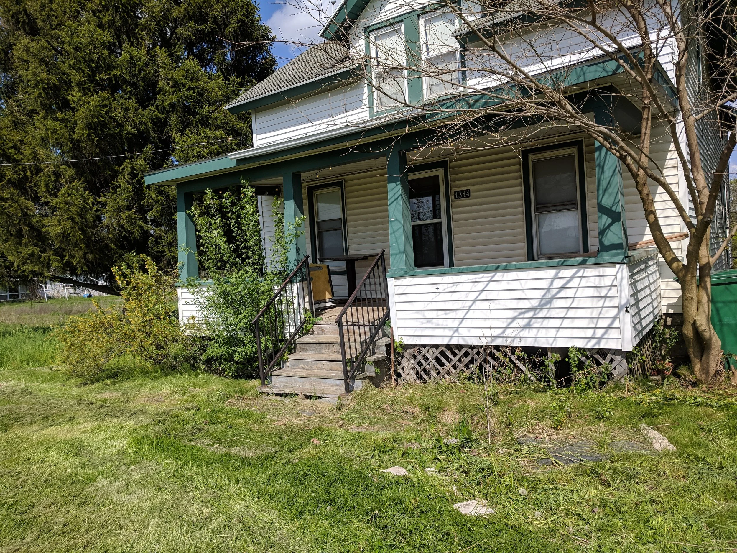 Front view of a two-story house with a green and white exterior, a small porch, and surrounded by greenery and trees.