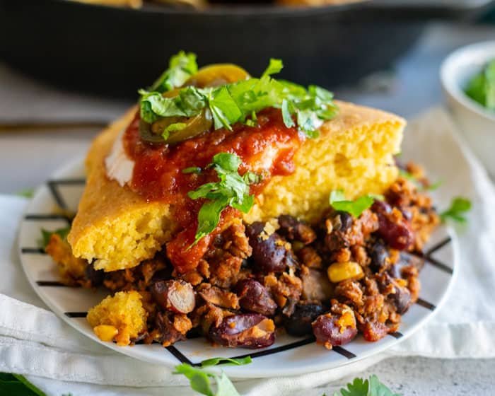A serving of cornbread topped with chili, salsa, jalapeños, and cilantro on a plate.