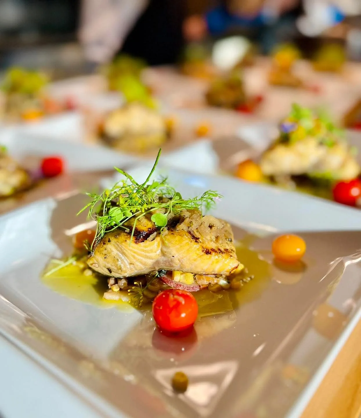 Grilled fish topped with herbs on a plate, with cherry tomatoes and sauce.