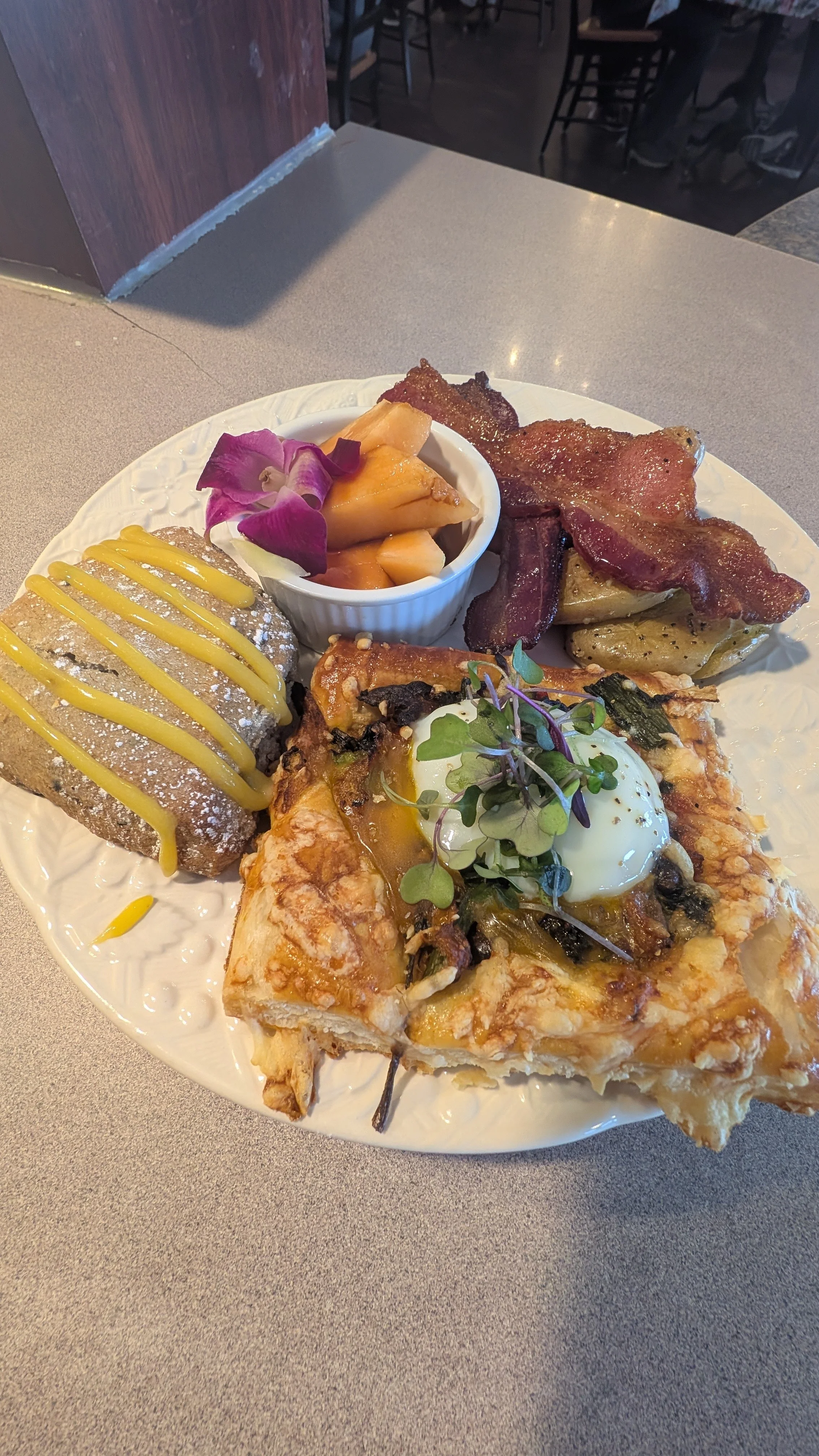Plate with bacon, roasted potatoes, a pastry topped with egg and greens, melon slices in a bowl with a flower garnish, and a scone drizzled with yellow icing.