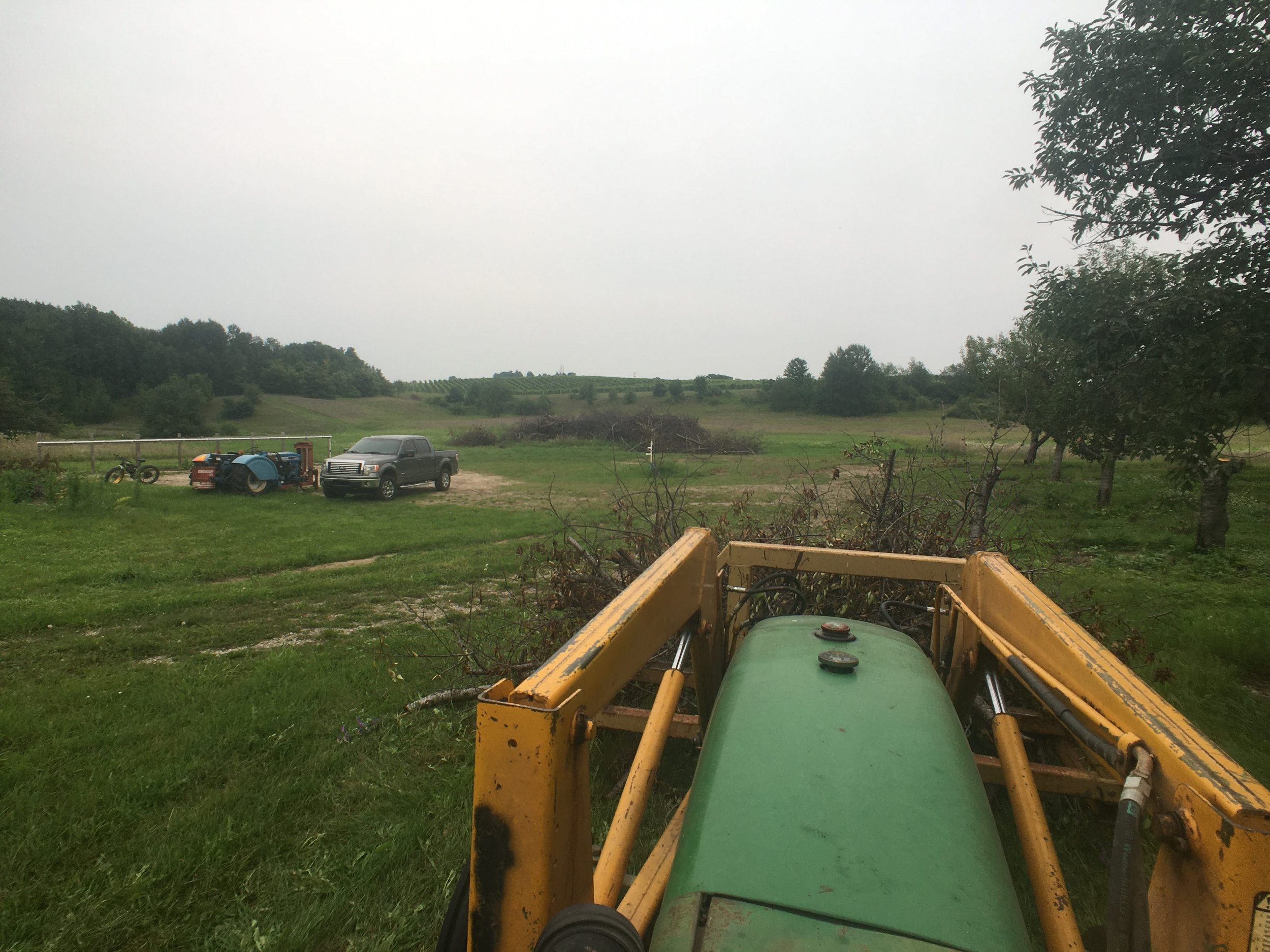 Tractor and truck parked in a rural grassy field with trees and rolling hills in the background on a cloudy day.