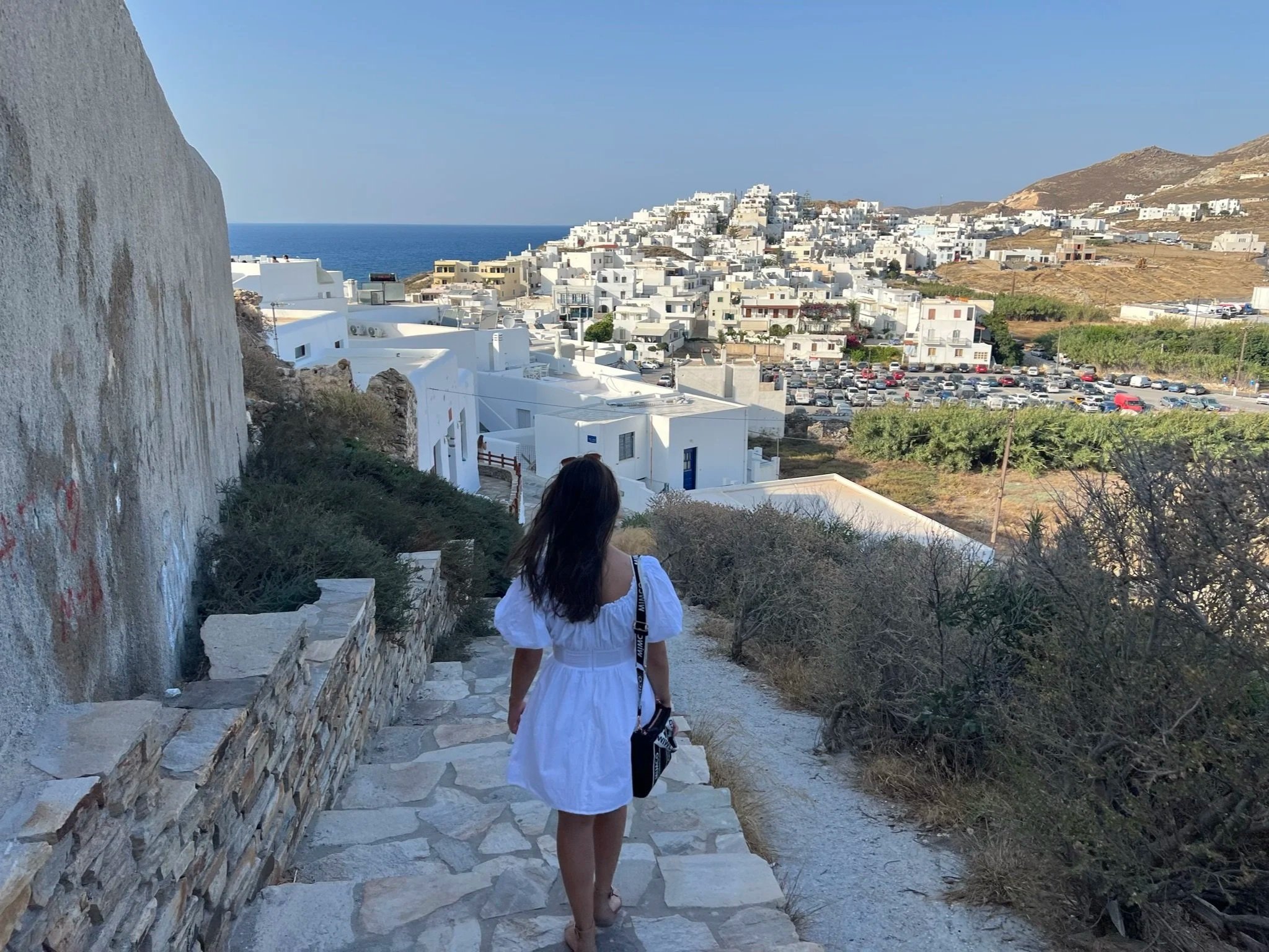 A woman in a white dress walking down stone stairs on a hillside, with white buildings and a parking lot in the distance, overlooking the sea under a clear blue sky.