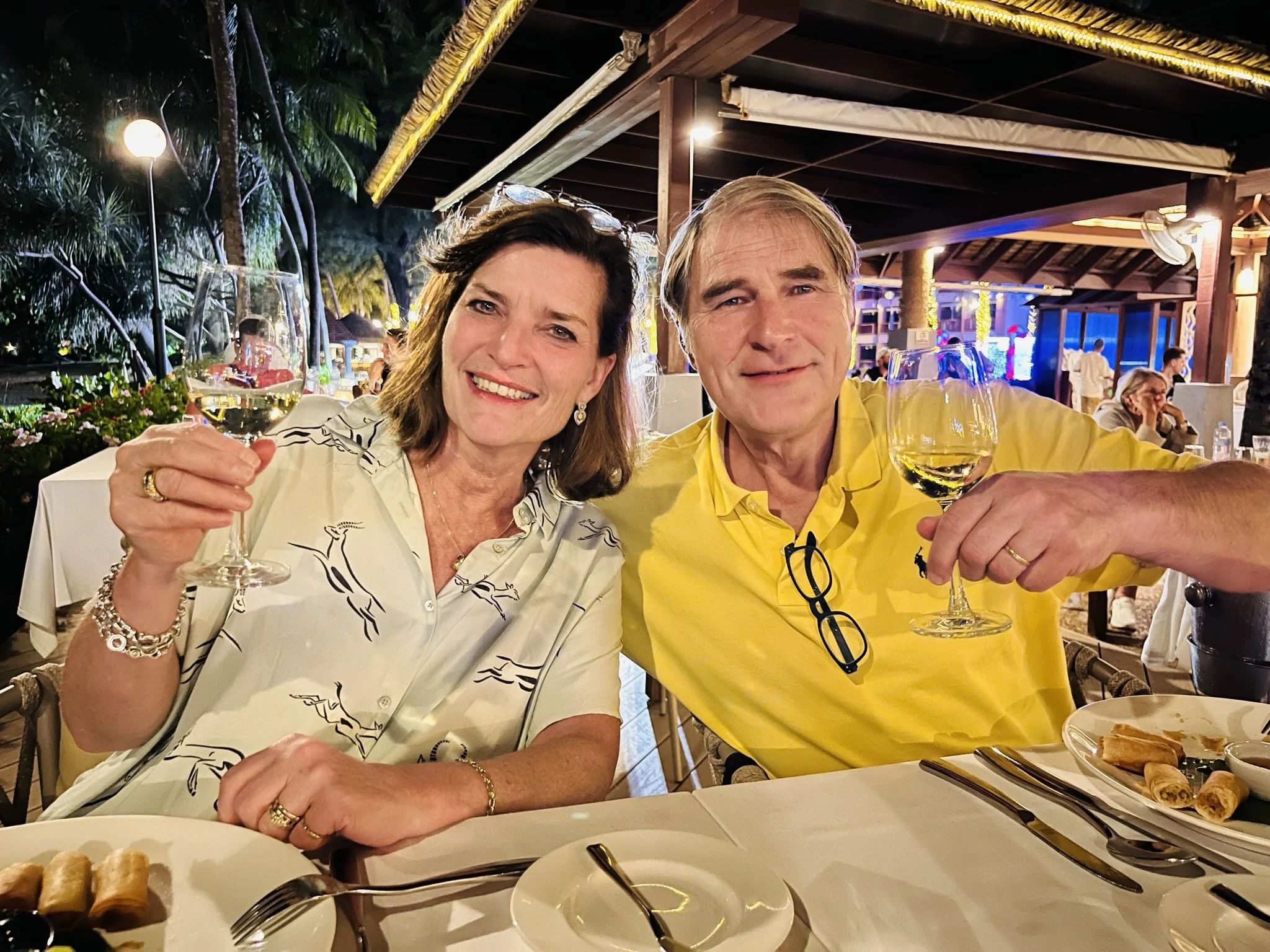 Twee mensen zitten aan een tafeltje in een restaurant of bar, beiden met een glas witte wijn omhoog en lachen.