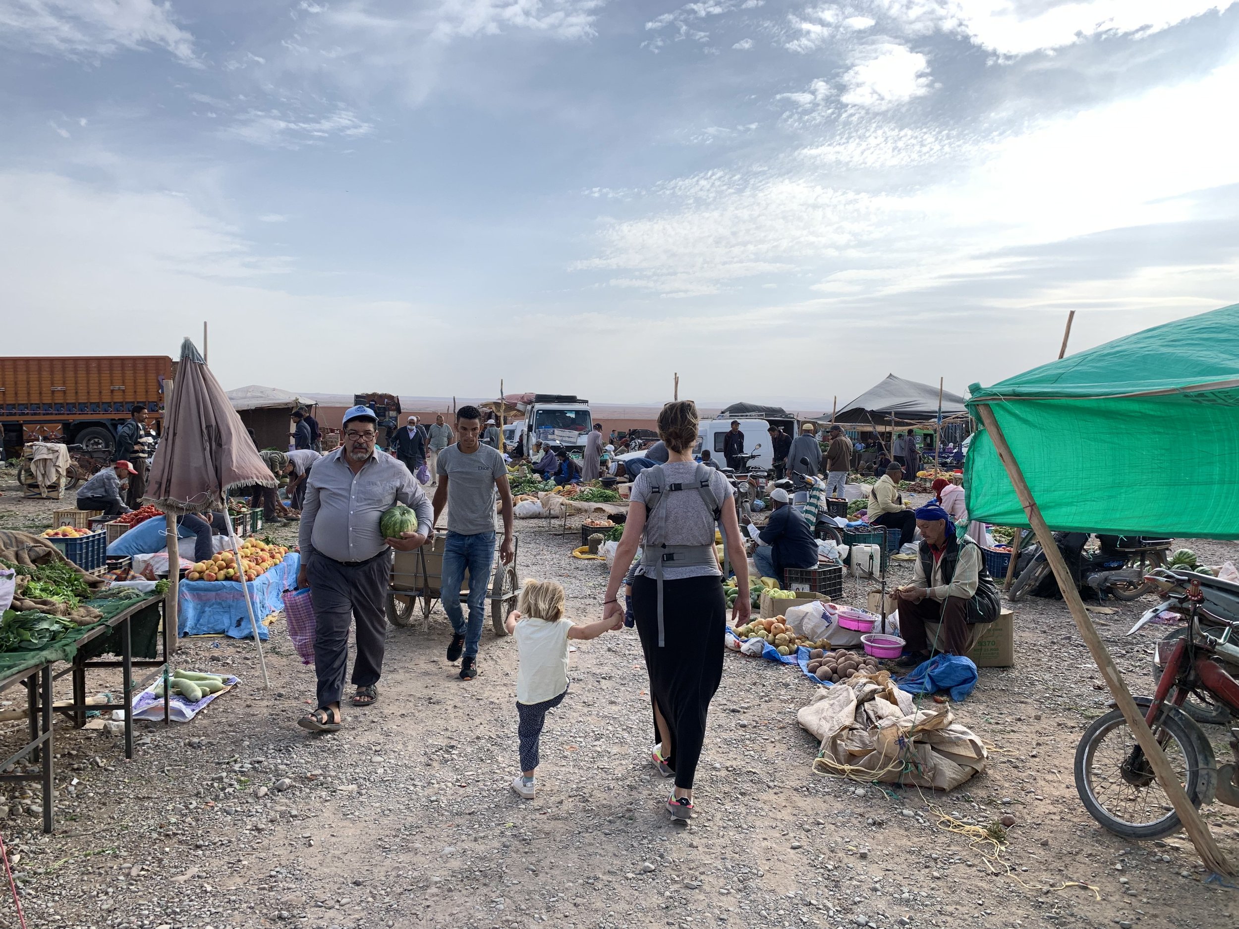 Een markt met verschillende mensen die bloemen, groenten en fruit verkopen. Er staan tenten en vrachtwagens op een open veld met een heldere, deels bewolkte lucht.