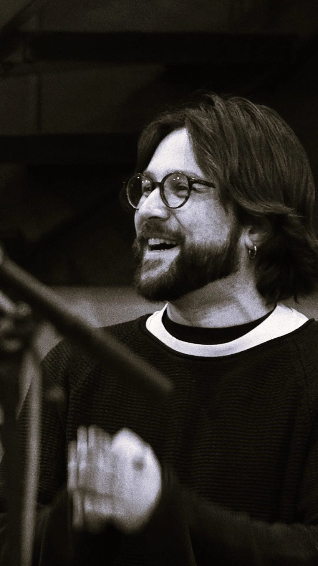 A man with shoulder-length hair, beard, and glasses singing or speaking, wearing a black and white shirt, in a recording studio.