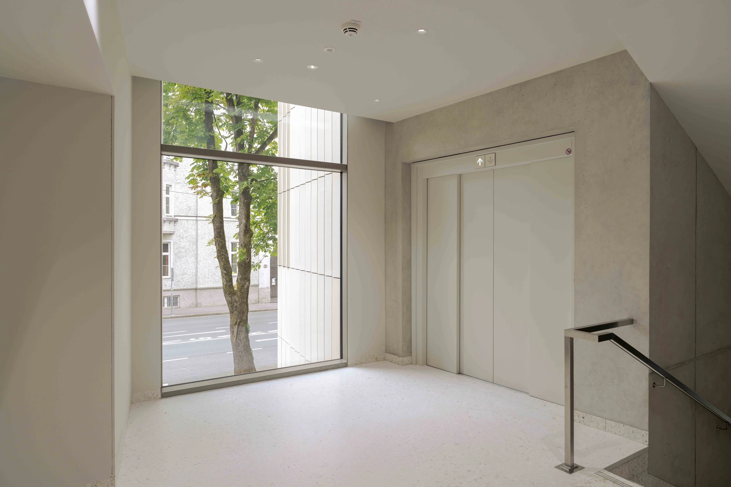 Ein heller Aufzugsschacht mit einer großen Fensterfront, durch die man eine Straße und Bäume sieht.