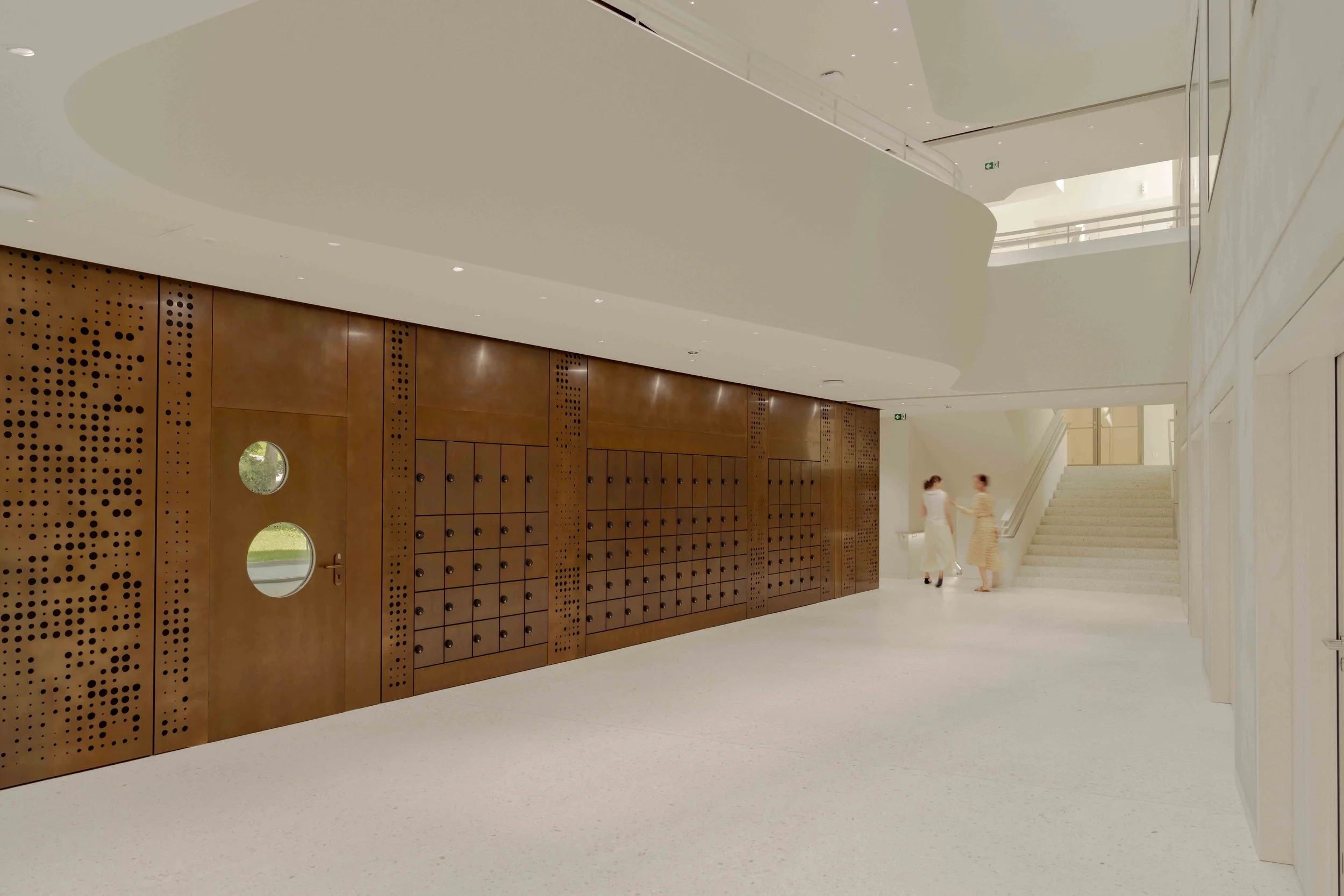 Moderner, heller Innenraum mit weißer Decke und Holz-Wand mit runden Türen, zwei Frauen gehen die Treppe hinauf.