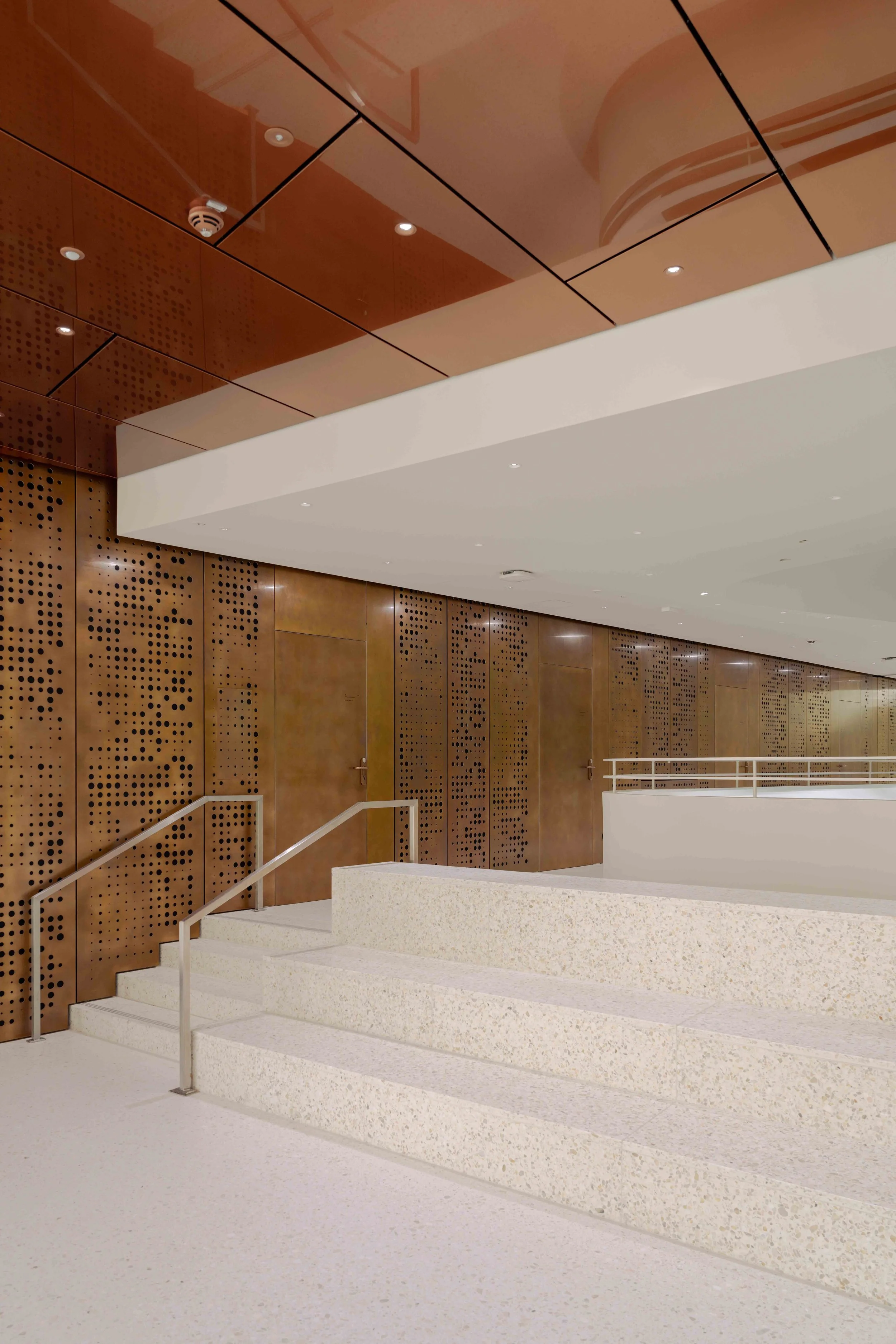 Ein moderner, heller Raum mit weißen Treppen, braunen Holzwänden mit perforierten Mustern und einer weißen Decke.