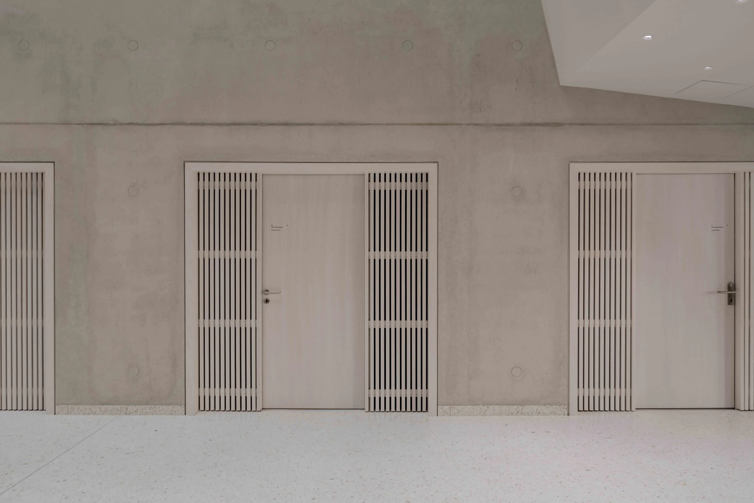 Drei geschlossene Türen mit vertikalen Holzlamellen in einer grauen, minimalistischen Wand, mit weißem Boden, moderner Innenarchitektur.