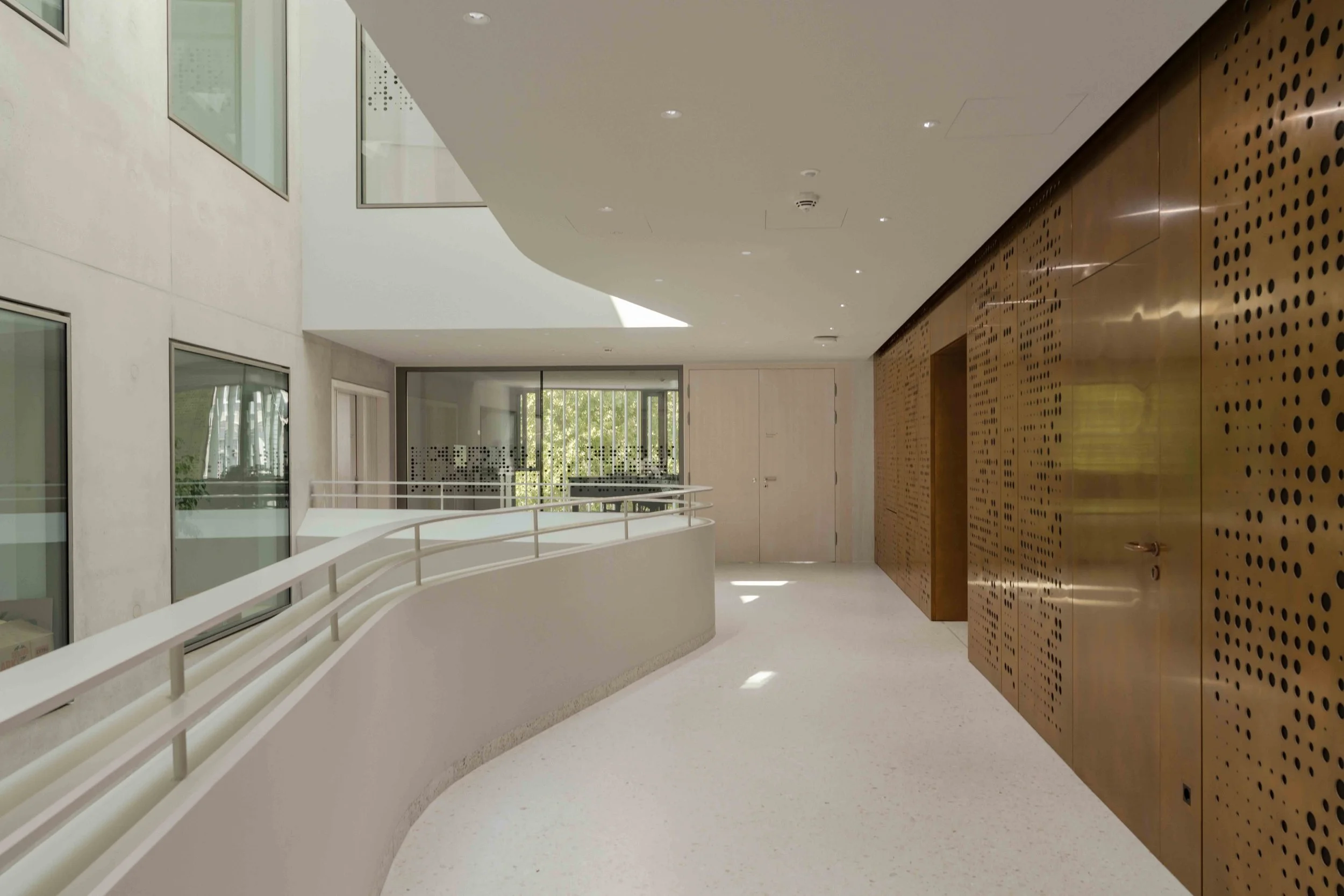 Innenansicht eines modernen Gebäudeflurs mit weißer Decke und Wänden, einer Garderobe aus Holz mit perforiertem Muster, großen Fenstern und einem Handlauf am Balkon.